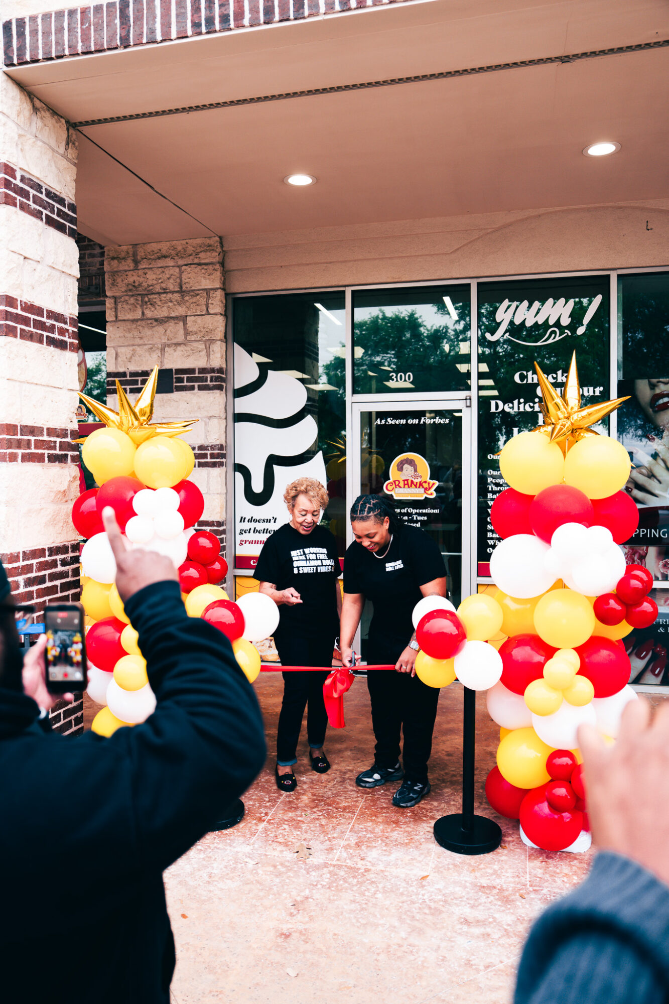 Two children stand behind a red ribbon at a balloon-decorated opening event outside a store, with people taking photos.