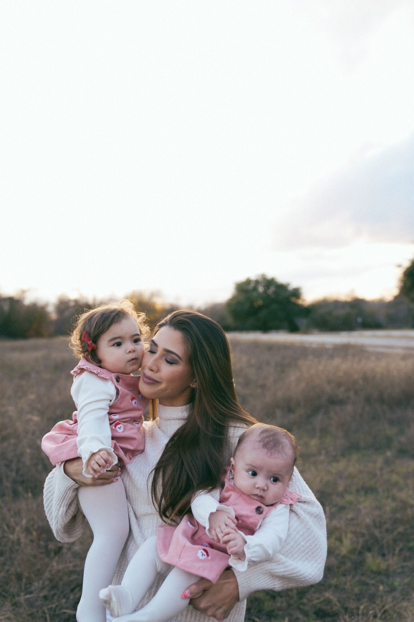 Woman holding two young children outdoors in a field, one child on her shoulder, the other in her arms, with a cloudy sky.