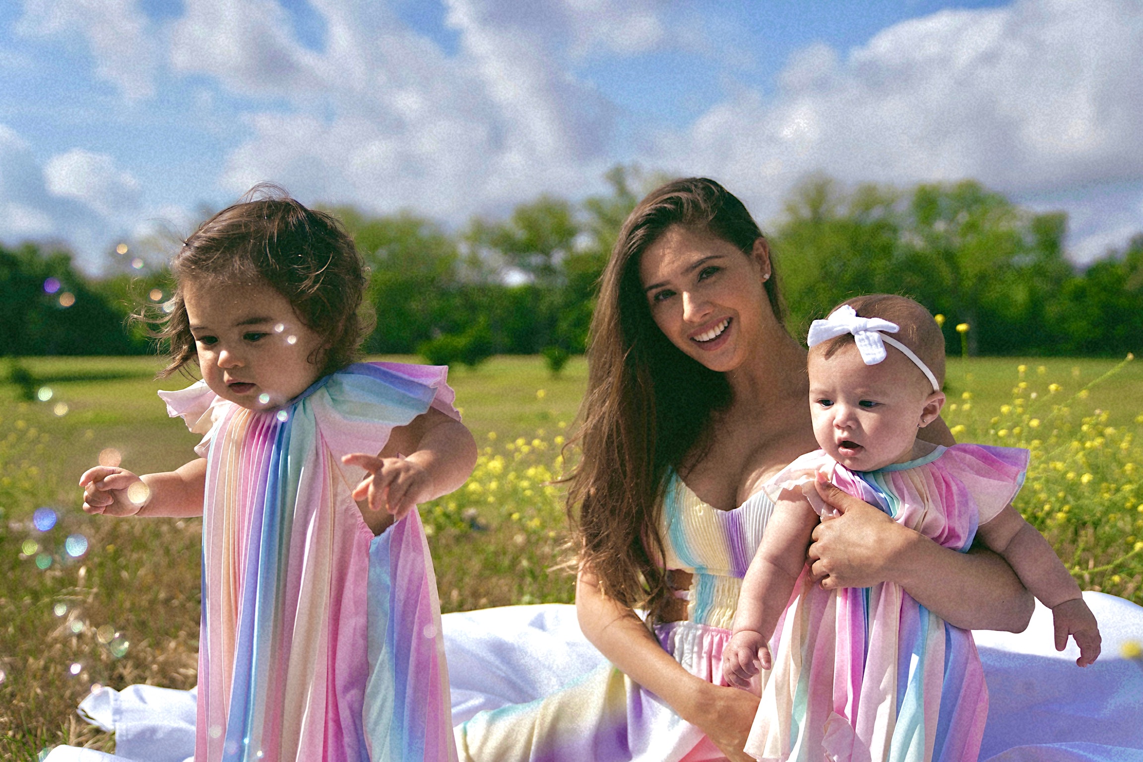 Woman holding a baby girl outdoors with a young girl nearby, all wearing colorful striped dresses, in a grassy field with trees and sky.
