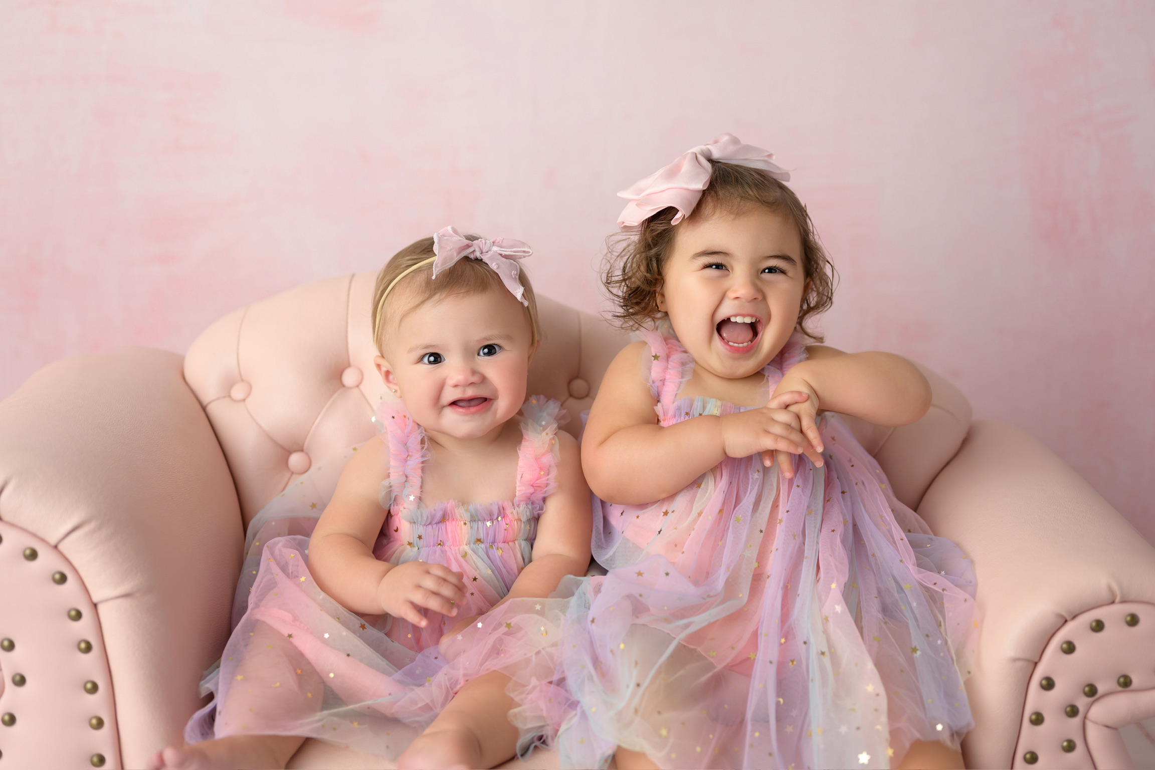 Two young girls in pastel dresses sitting on a beige sofa, smiling and laughing, with pink background.