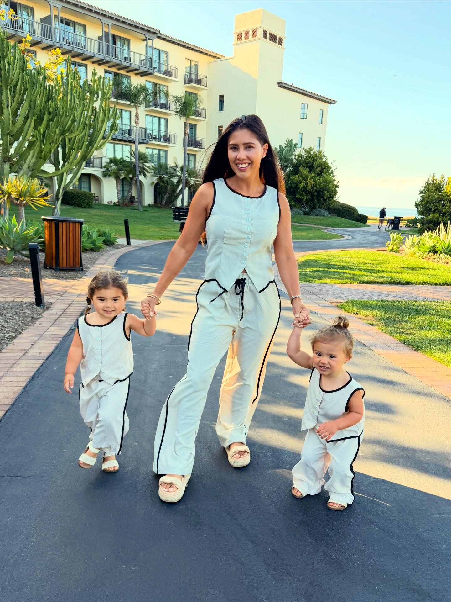 Woman walking with two young children on a paved path in a park, with a building and greenery in the background.