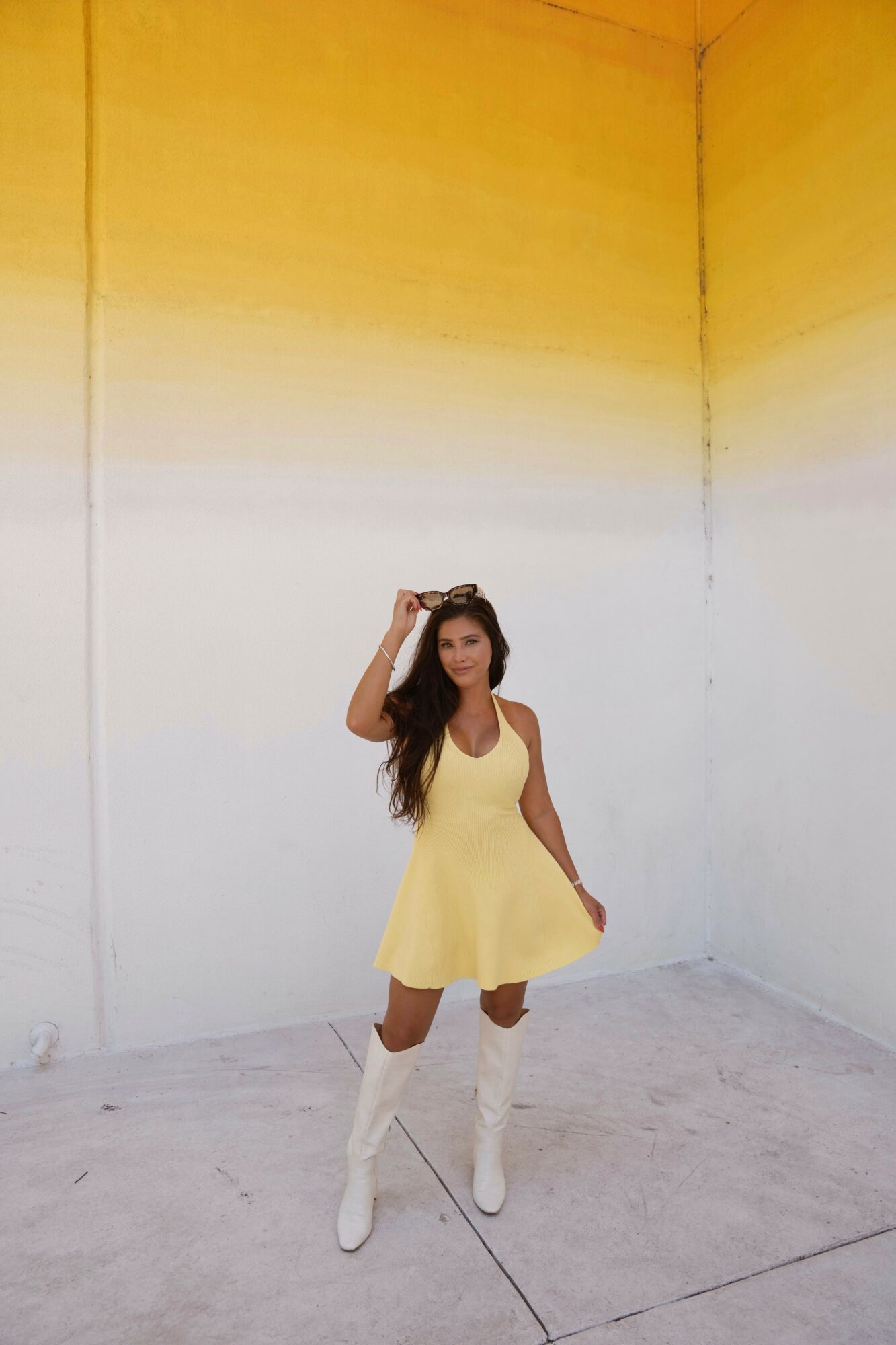 Young woman in yellow dress and white boots standing in corner with yellow and white walls, smiling, hand on head.