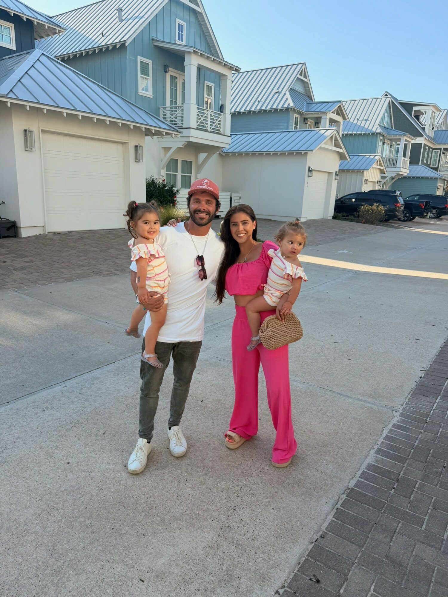 A man and woman each holding a child, standing on a sidewalk in front of blue houses, smiling at the camera.