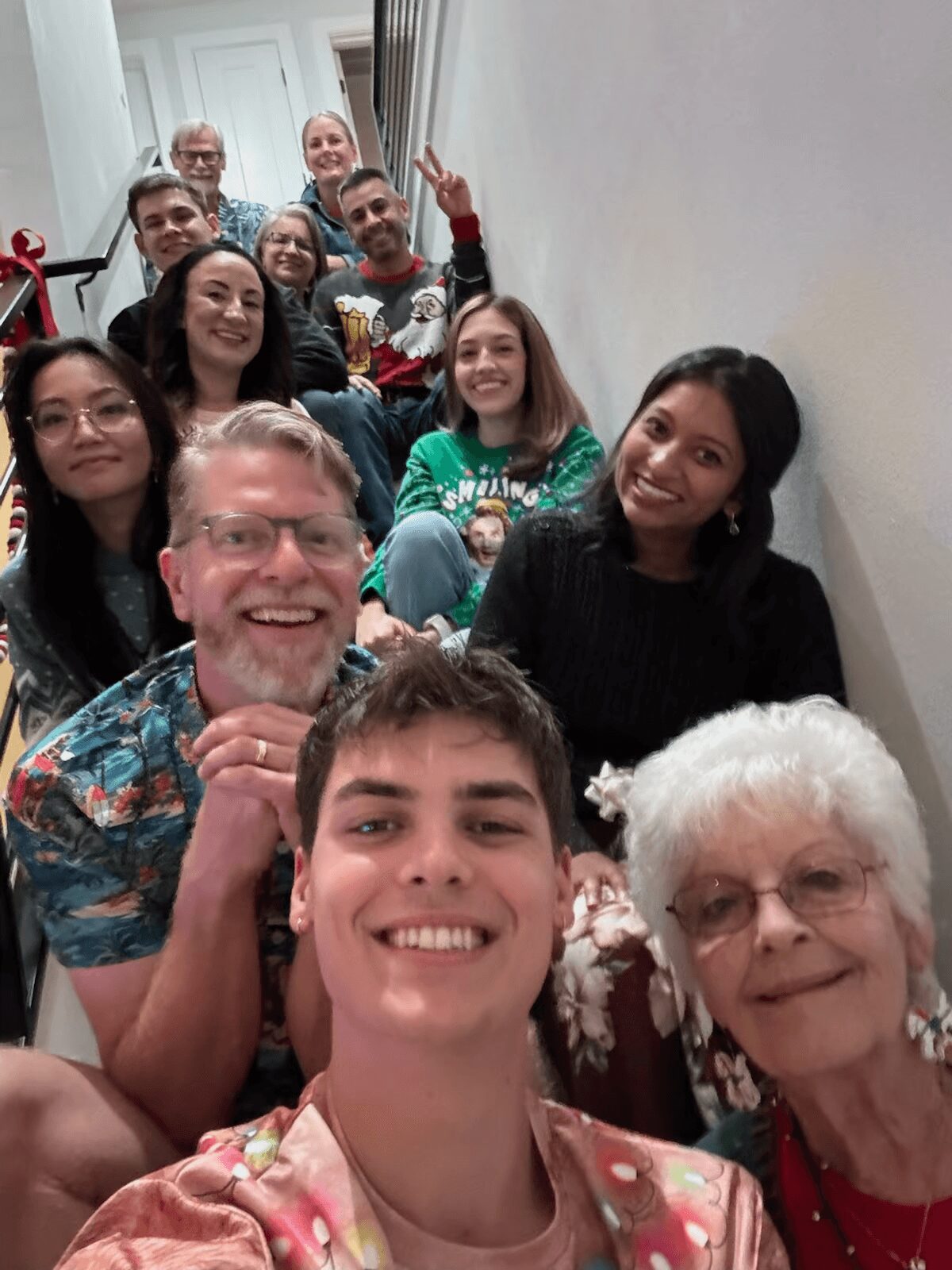 Group of people, including young and elderly, smiling and sitting together indoors, some making peace signs, in a narrow hallway.