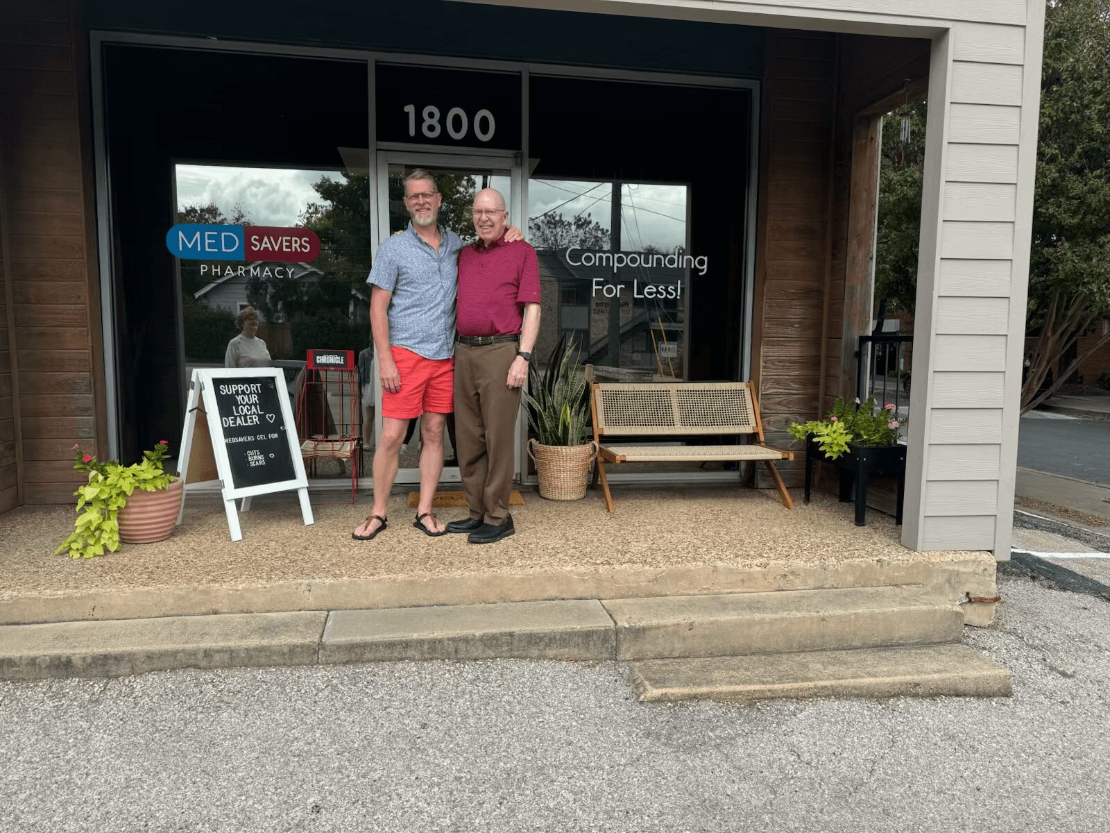 Two men stand outside a storefront with a sign reading 'Compounding For Less!' and a window displaying pharmacy-related signs.
