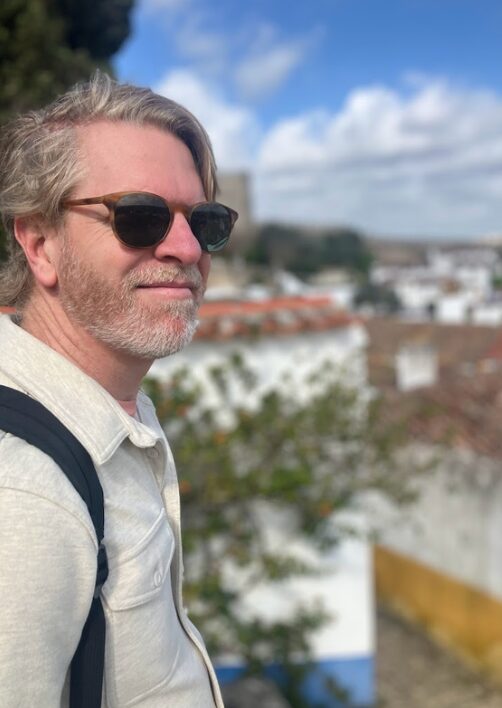 Man with gray hair and beard wearing sunglasses and a light-colored shirt outdoors, with a cityscape and blue sky in background.