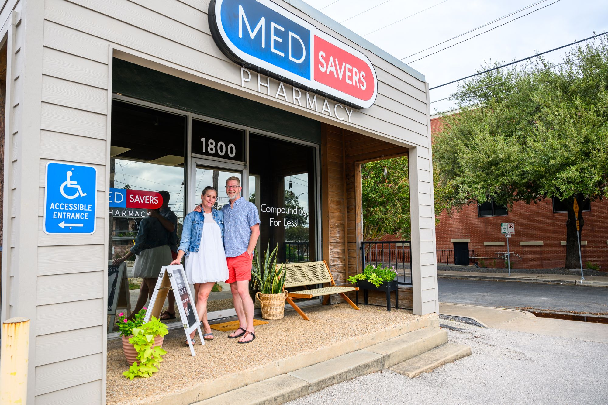 Two people stand outside a pharmacy with a blue and red sign, near potted plants and a bench, on a sidewalk.