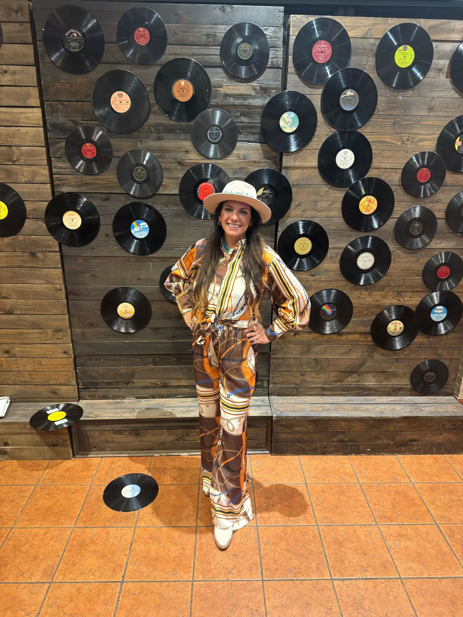 Person standing against a wall decorated with vinyl records, wearing a colorful patterned outfit, with a shadow on the tiled floor.