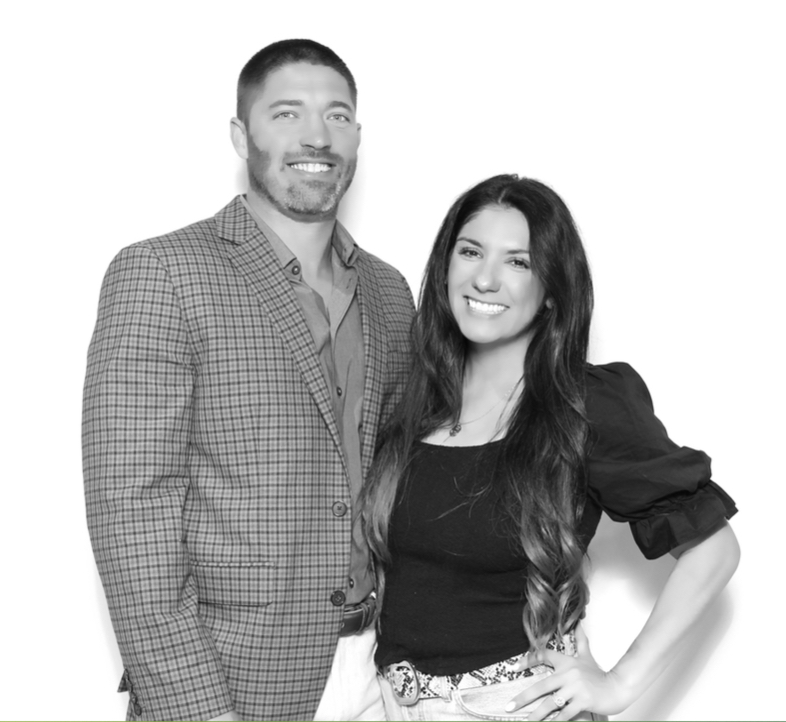 Two people, a man and a woman, smiling and standing close together, with the woman having long dark hair and the man wearing a checked blazer.