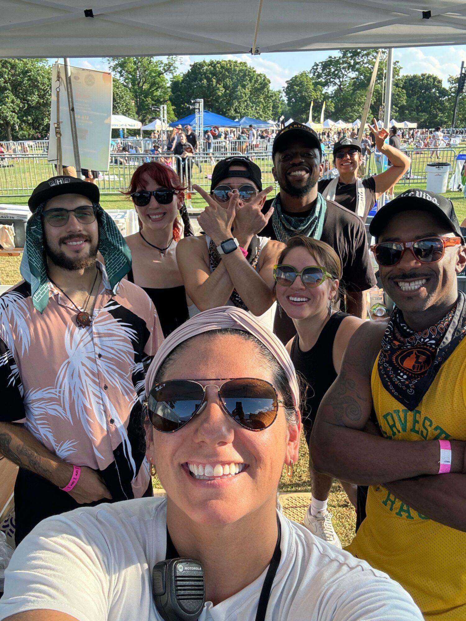 Group of six people smiling outdoors at a festival or event, some wearing sunglasses, with tents and trees in background.