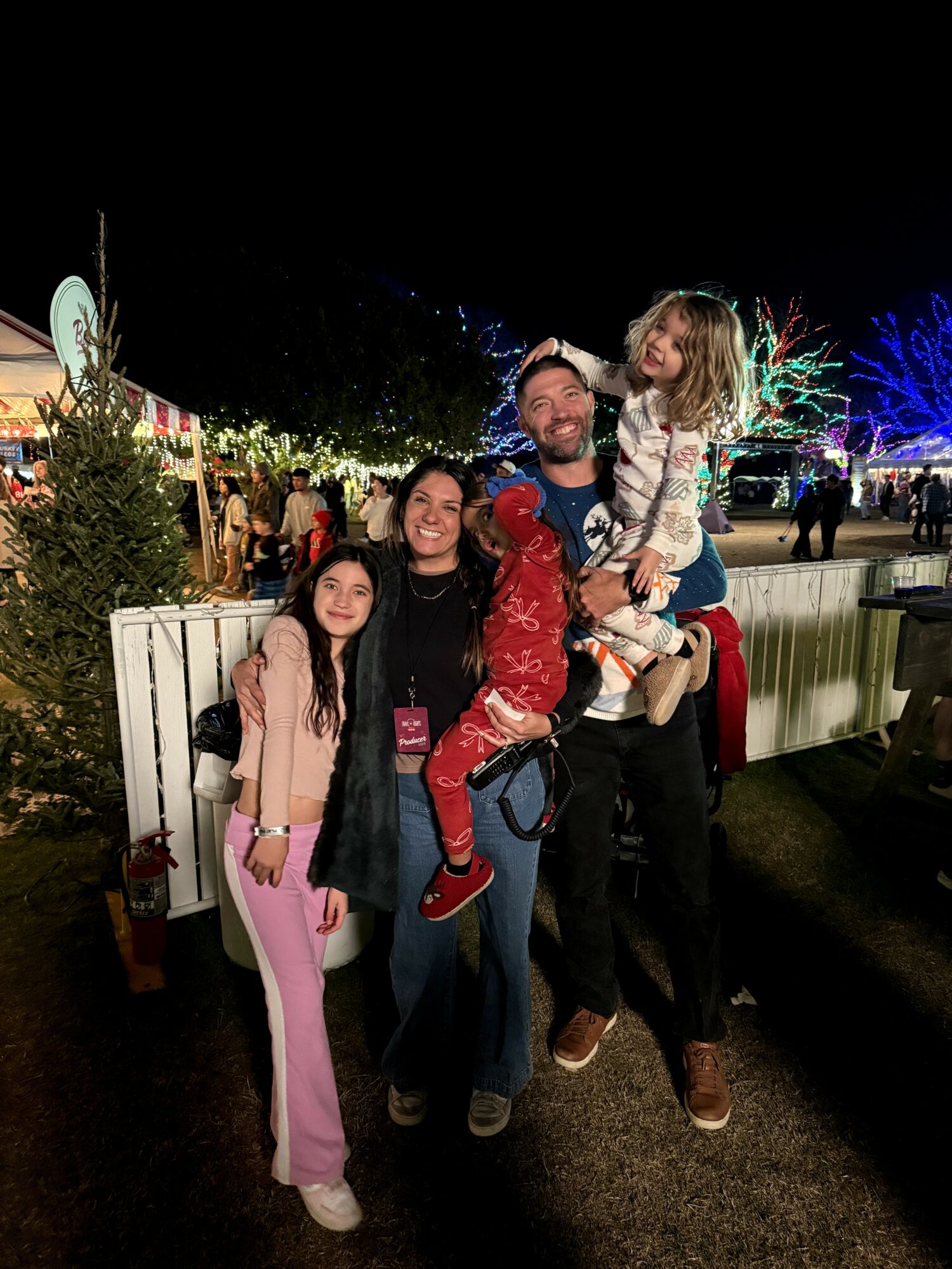 Group of four people, two adults and two children, smiling outdoors at night with festive lights and trees in background.