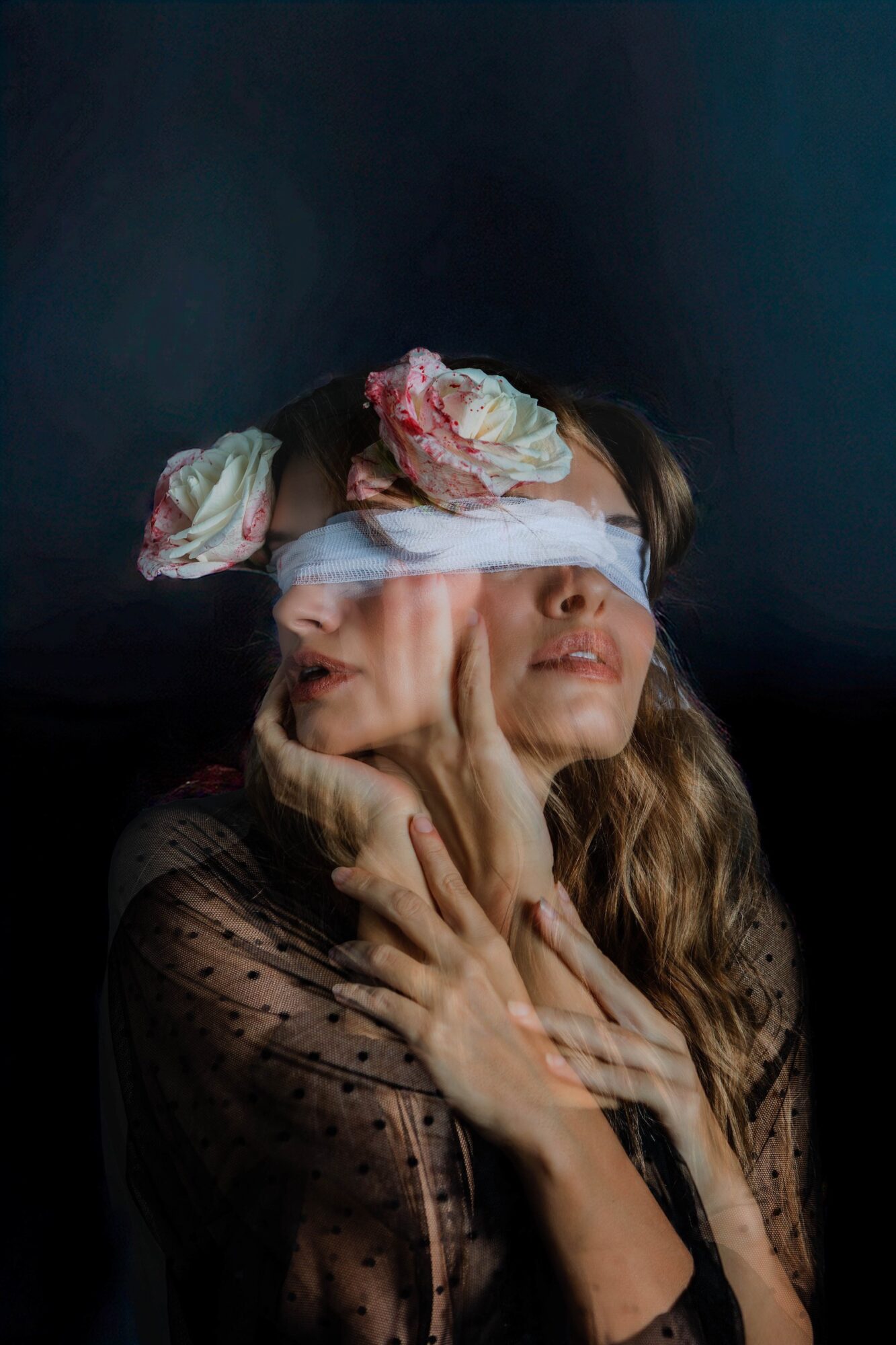 A woman with long hair has a cloth over her eyes, with flowers on her head, and her hands on her face.
