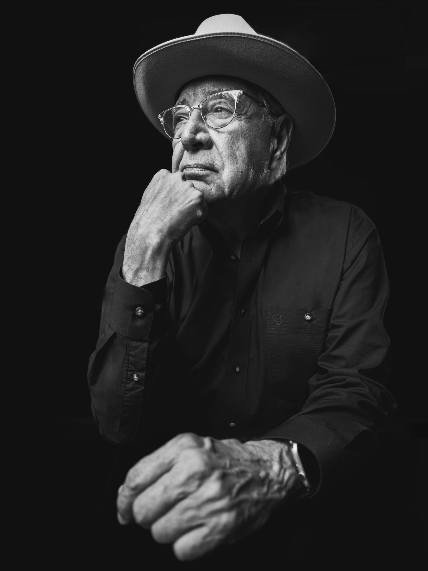 Elderly person wearing glasses and a wide-brimmed hat, resting chin on hand, in black and white.