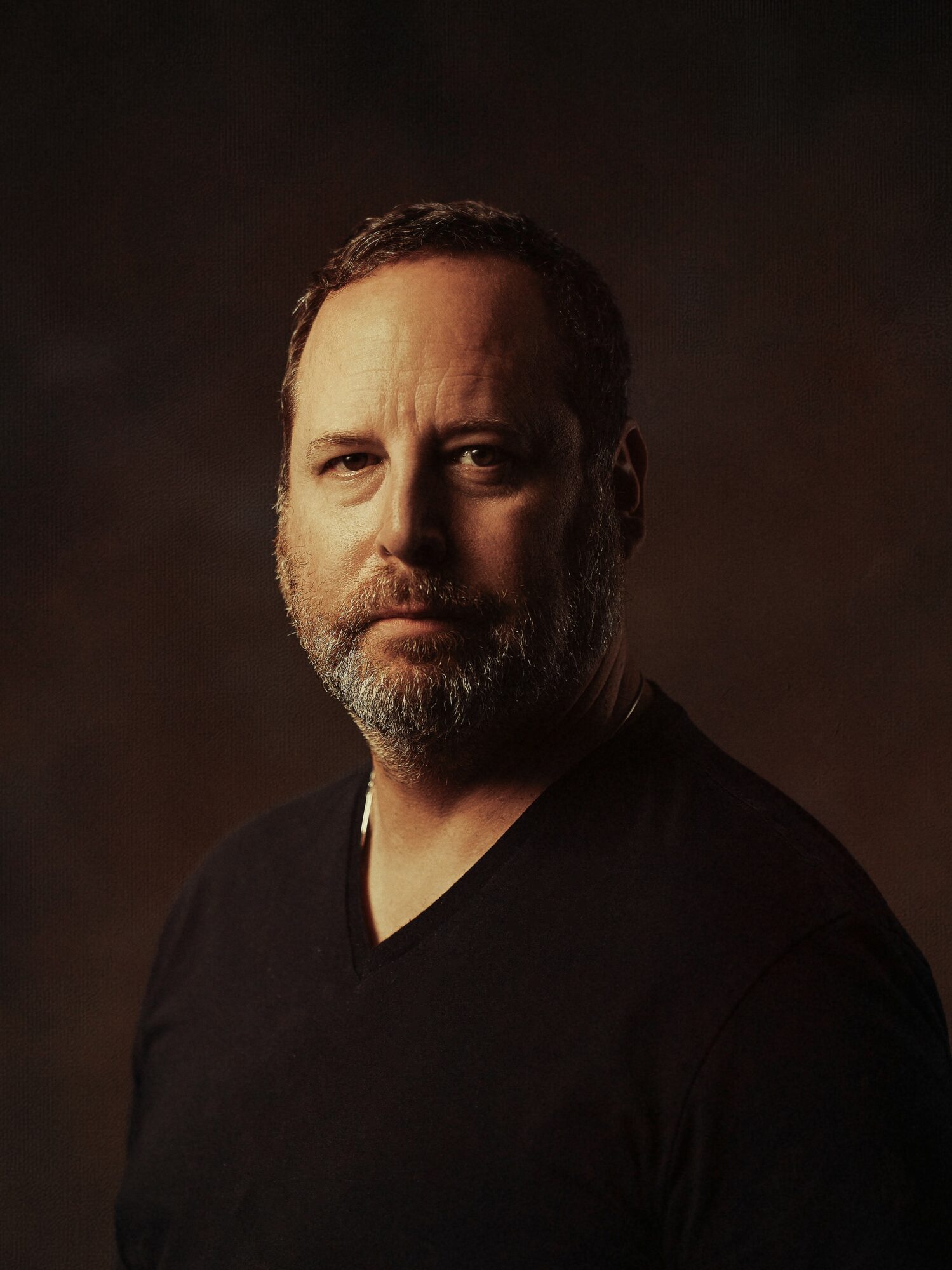 Portrait of a middle-aged man with a beard, wearing a black shirt, looking at the camera against a dark background.