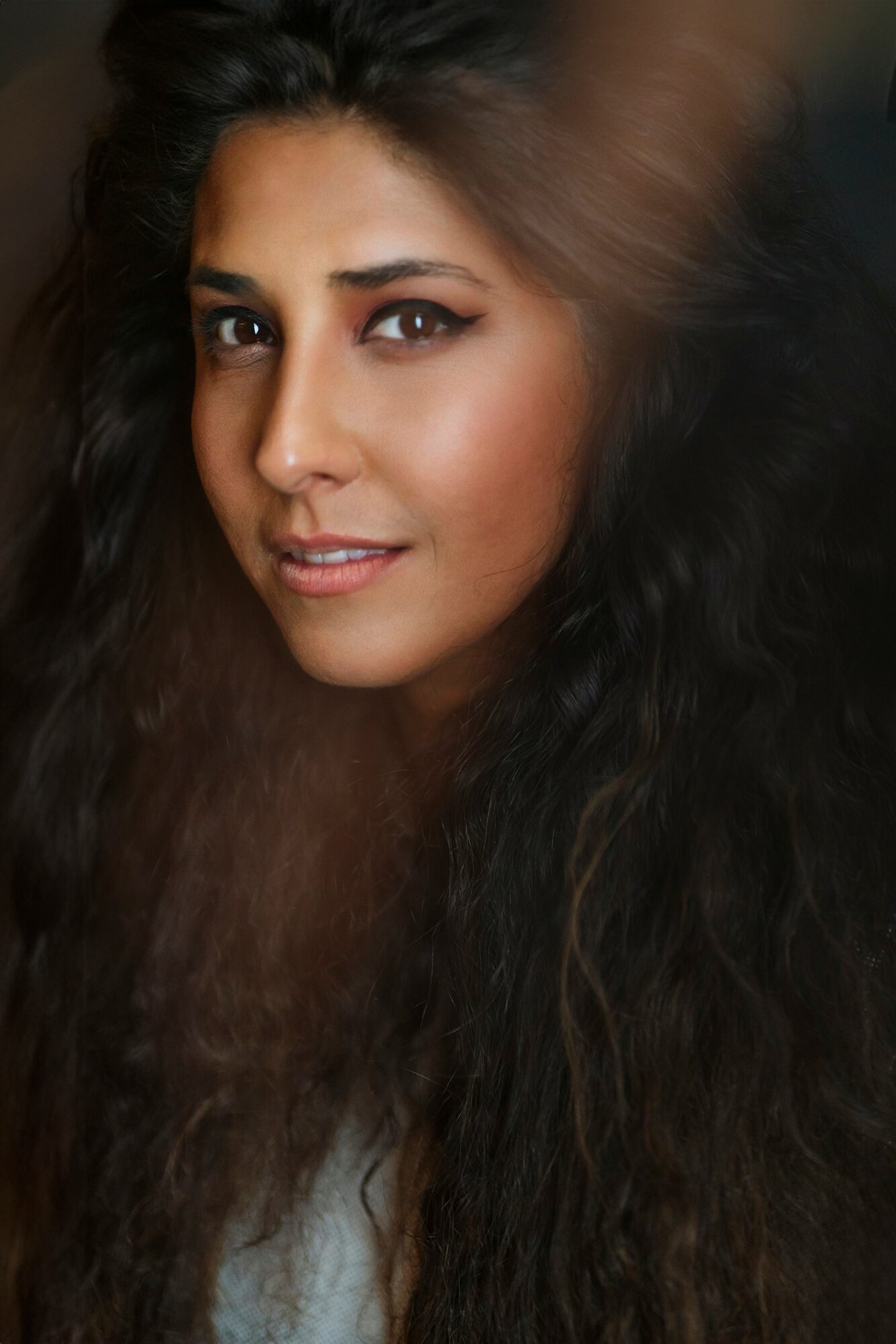 Close-up of a woman with long, dark, wavy hair and makeup, looking at the camera with a slight smile.