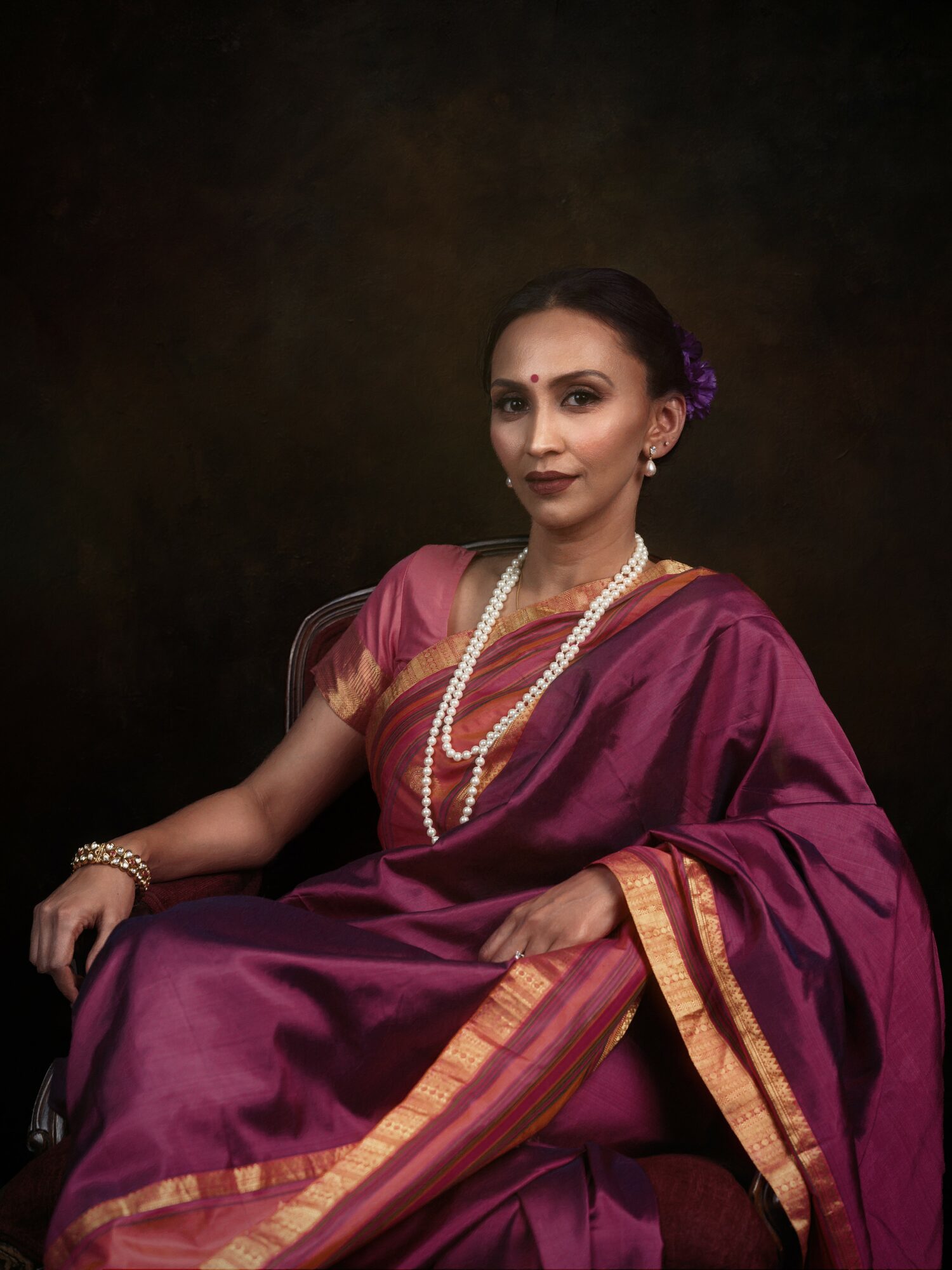 Woman in traditional attire with jewelry, sitting against a dark background.