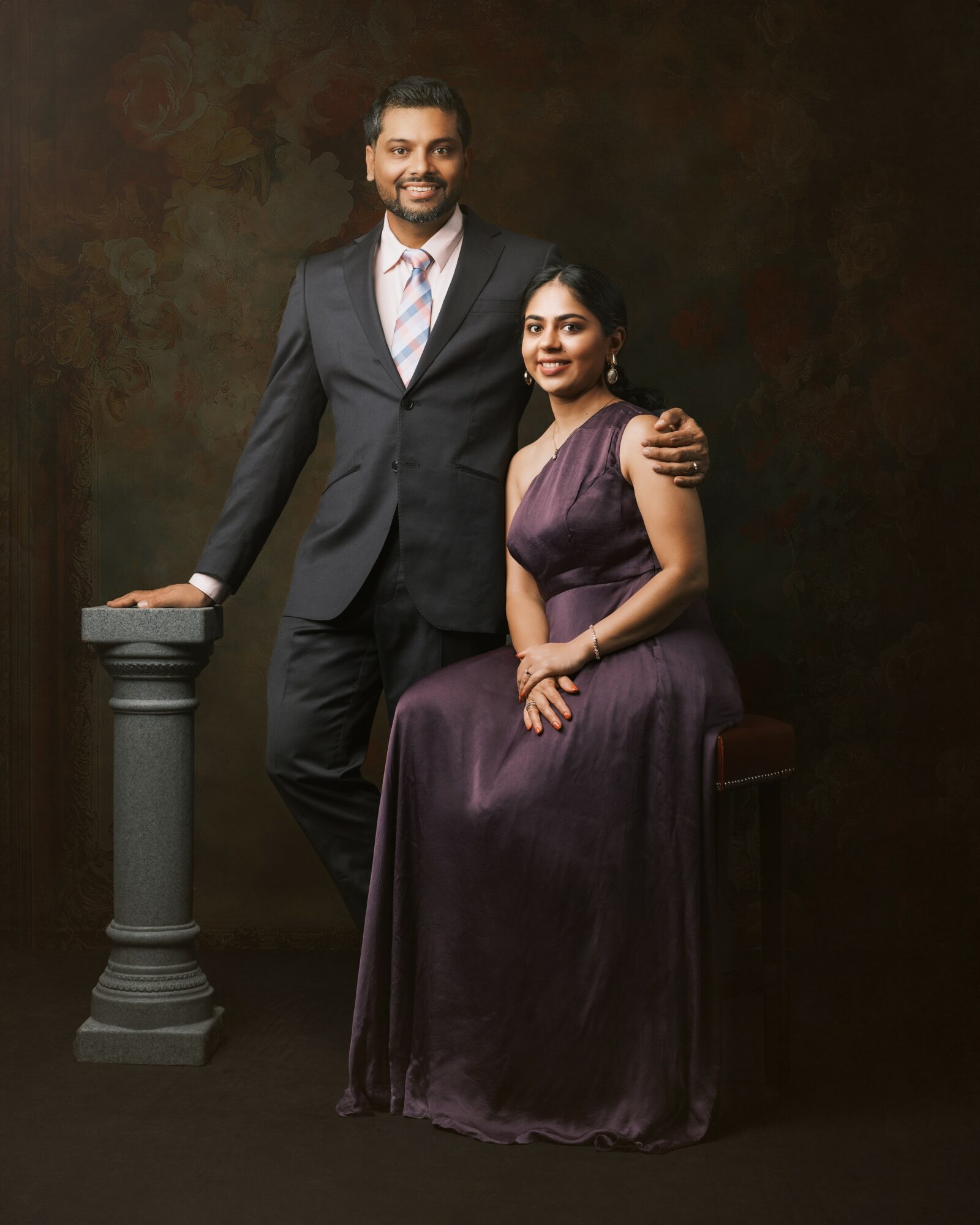 A man in a suit and a woman in a purple dress pose together against a dark background.
