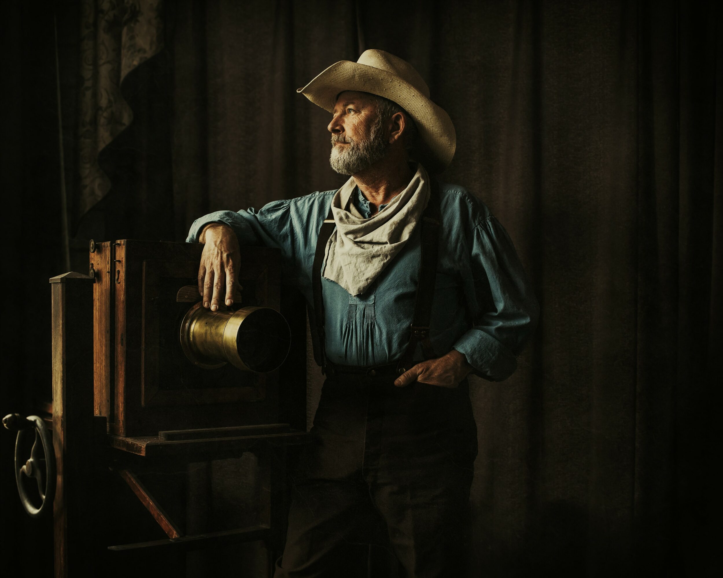 Man in cowboy hat and scarf, standing next to a vintage coin-operated device, with dark curtains in background.