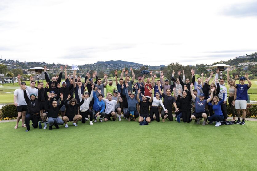 Group of people outdoors on grass, raising hands, with hills and cloudy sky in background.