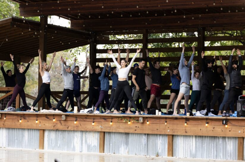 Group of people participating in an outdoor dance or exercise class under a wooden shelter, raising their arms.