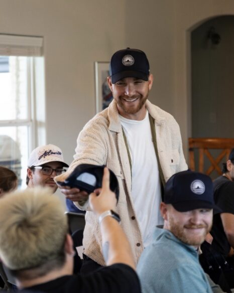 Smiling man in beige jacket and black cap holding a microphone, surrounded by seated people in a room with a window.