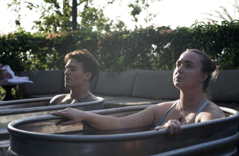 Two people sitting in a hot tub outdoors, with greenery and a hedge in the background, during daytime.