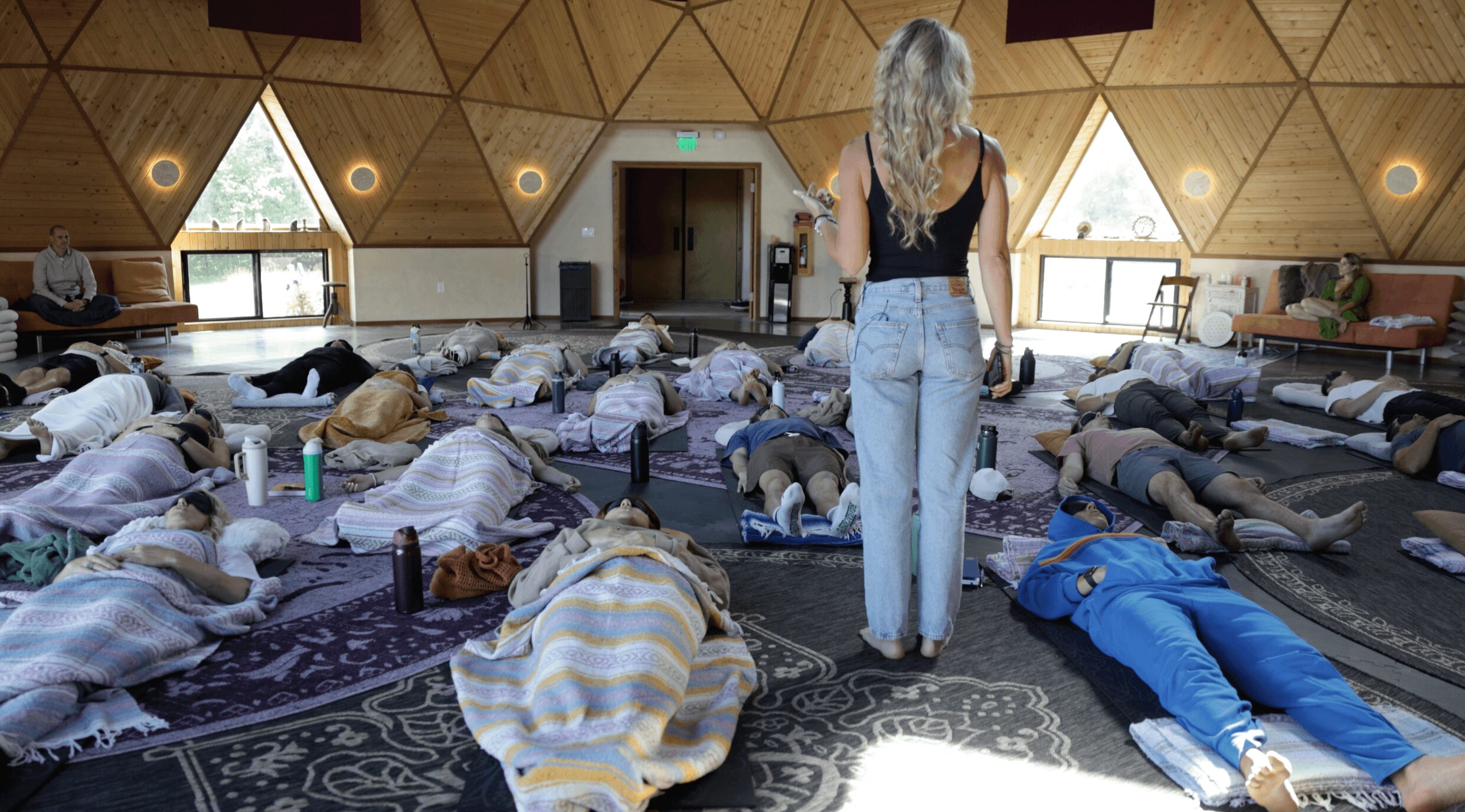 Group of people lying on mats in a spacious room with wooden walls and large windows, a woman standing in the center.