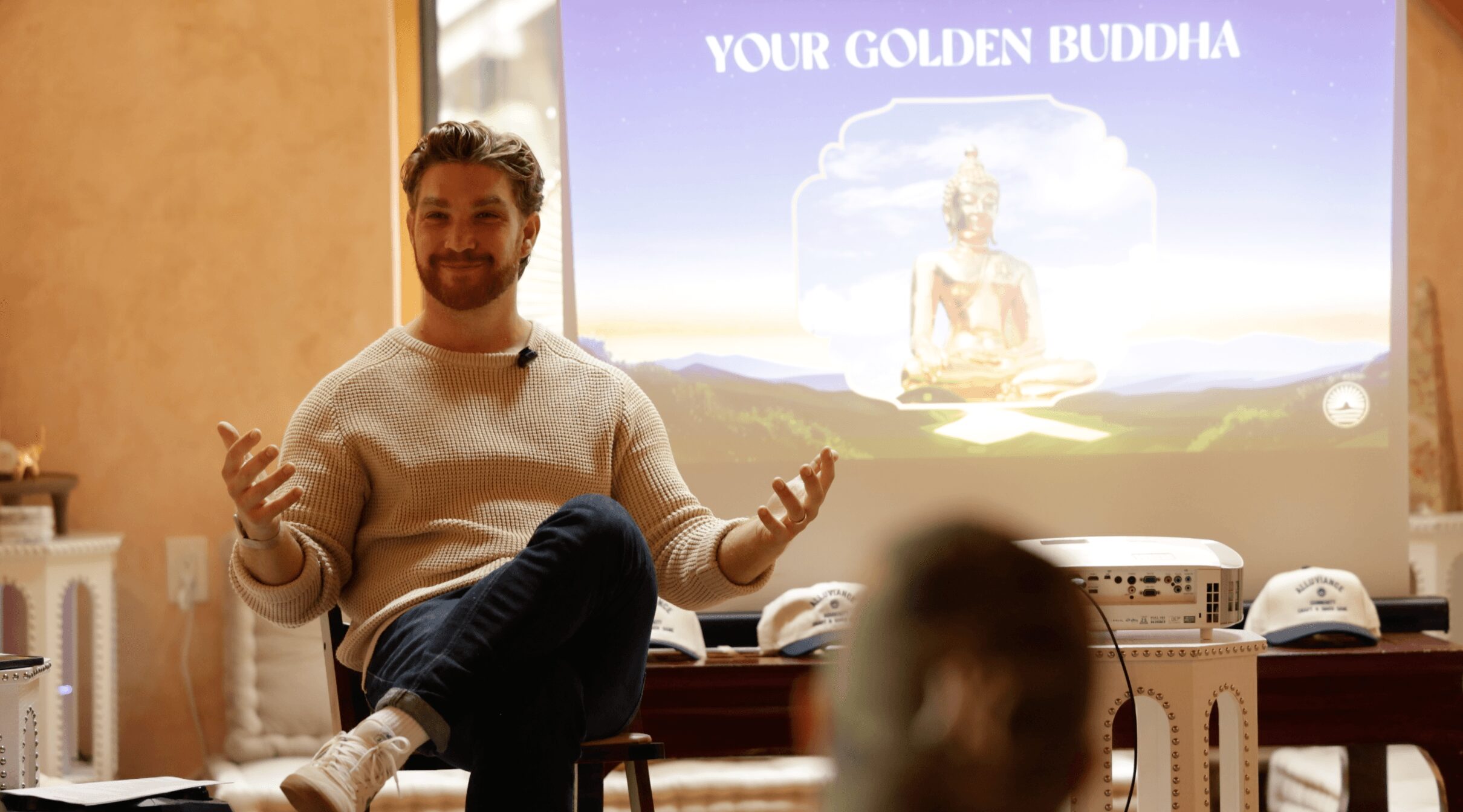 Person sitting on a chair in front of a presentation screen, gesturing with hands, with a blurred audience in foreground.