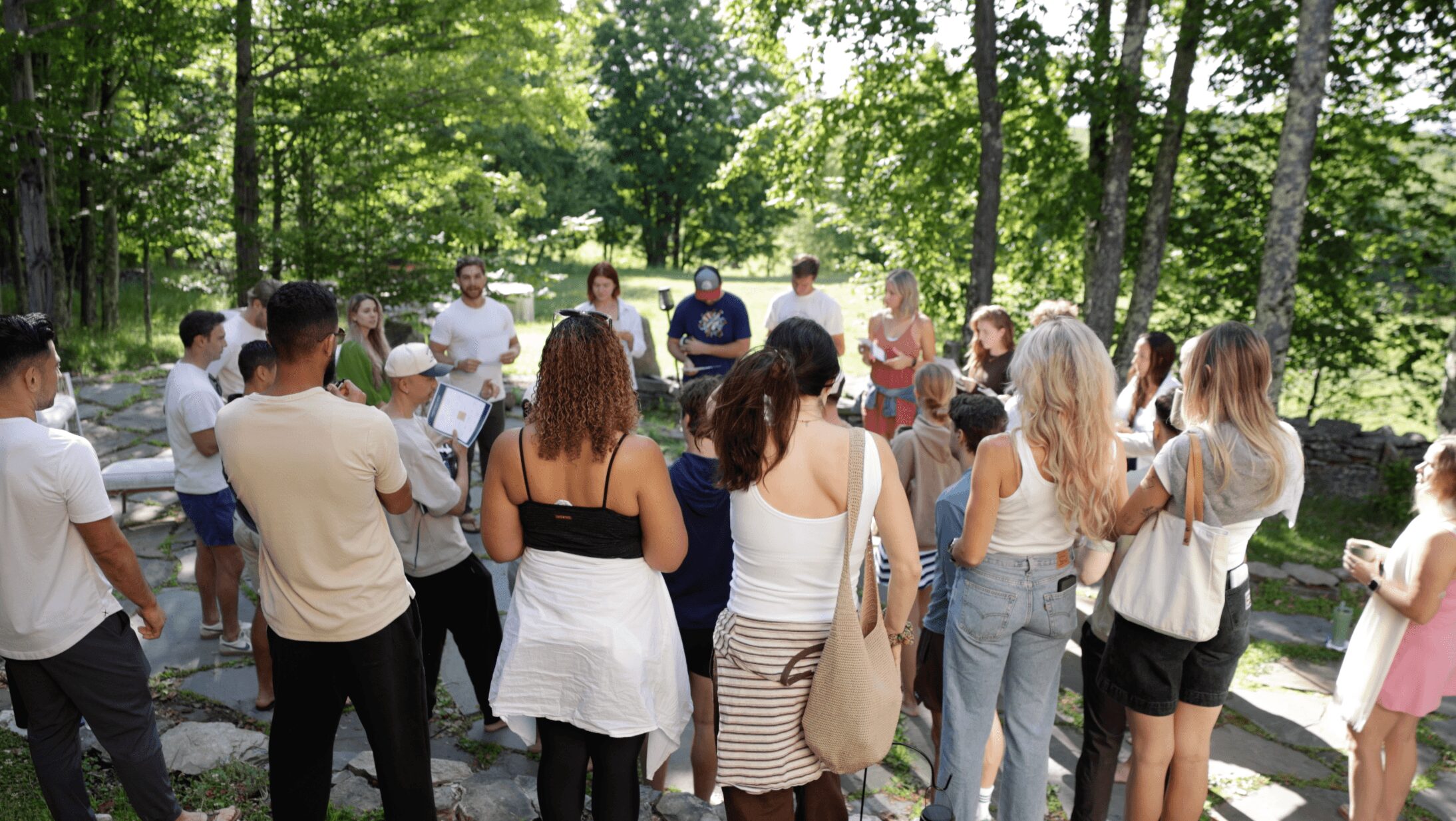 Group of people gathered outdoors among trees, some holding papers, in a sunny setting with lush greenery.