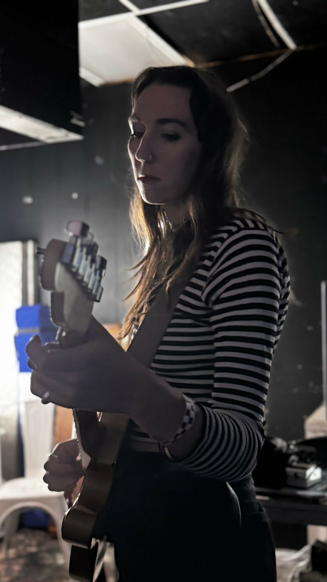 Woman with long hair in striped shirt holding a guitar, dimly lit background.