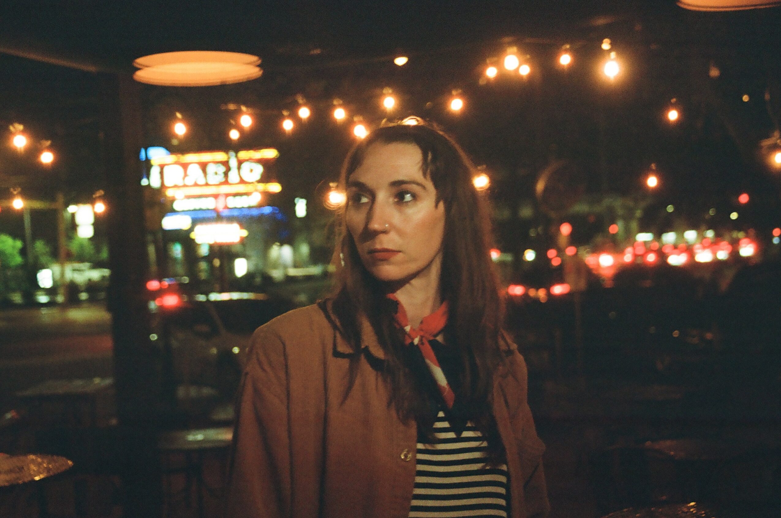 Woman with long dark hair standing outdoors at night with city lights and streetlights in background.