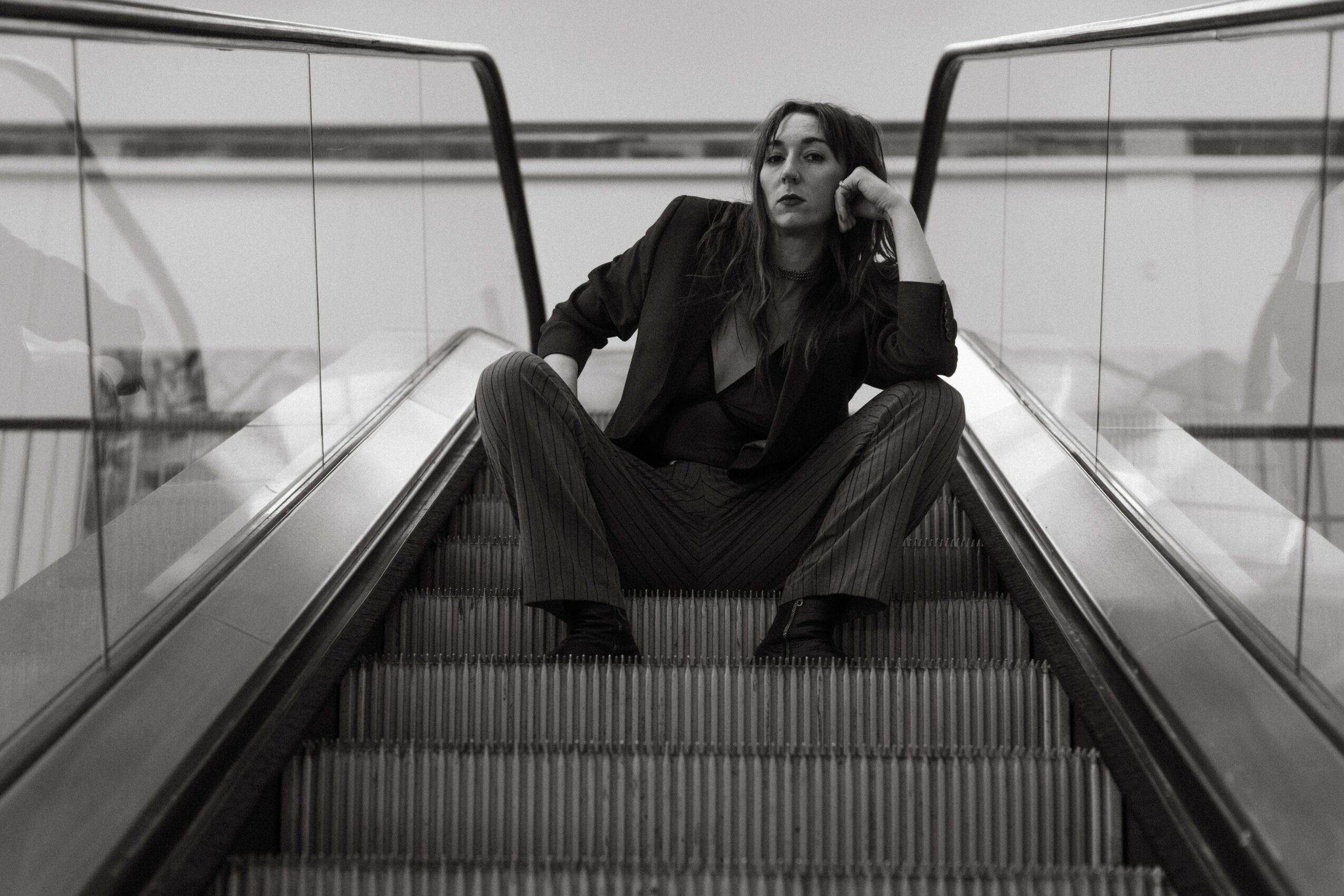Woman sitting on escalator steps, resting her head on her hand, in black and white, with glass railings on sides.
