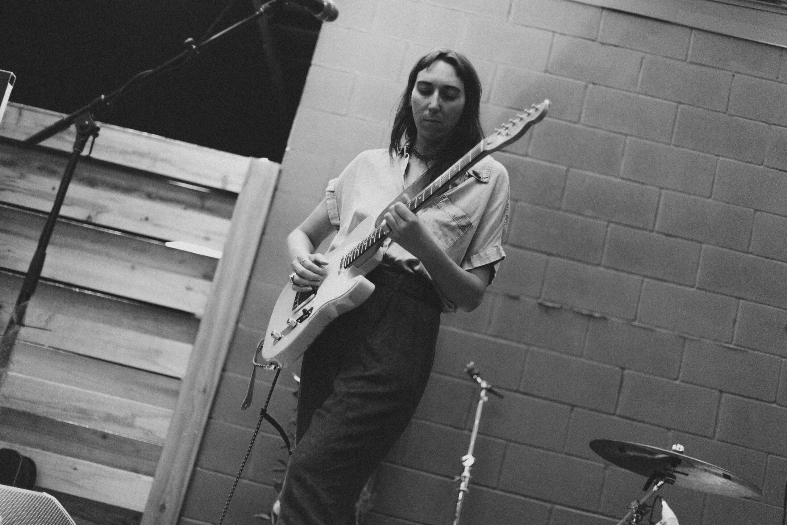 A woman playing an electric guitar on stage, with a brick wall background, in black and white.