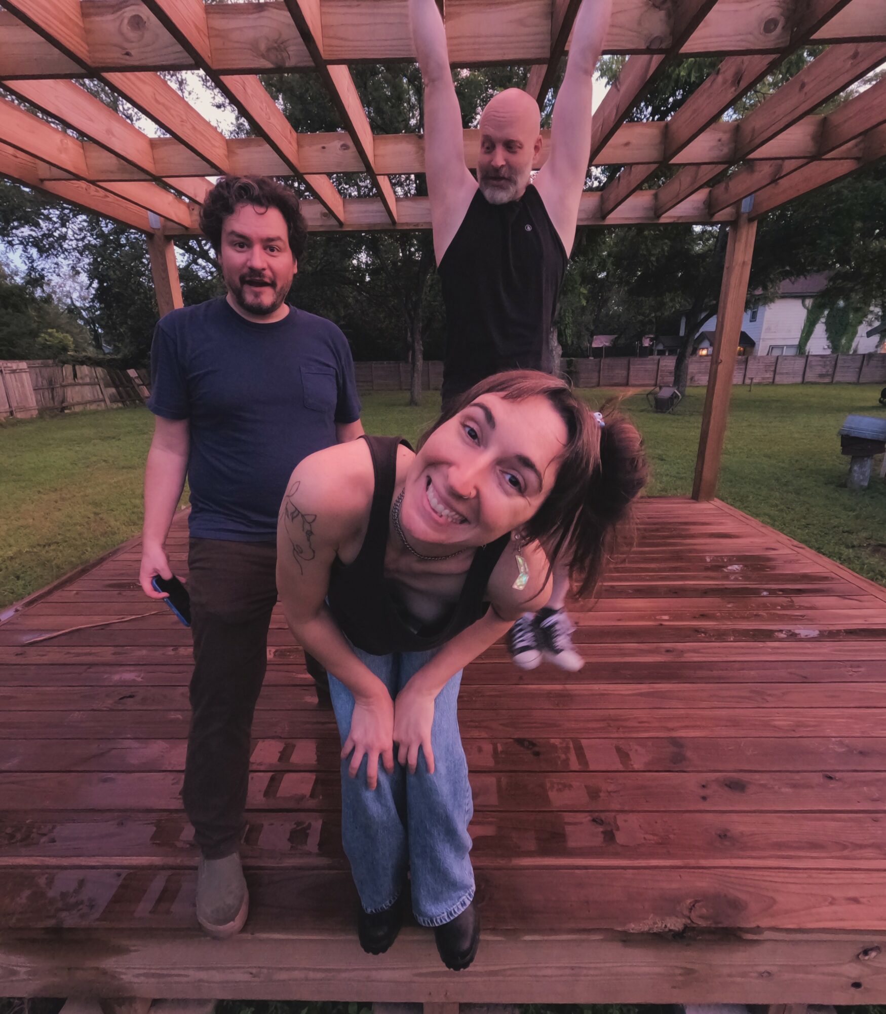 Three people on a wooden deck outdoors, one hanging from a wooden structure, smiling and posing for the photo.