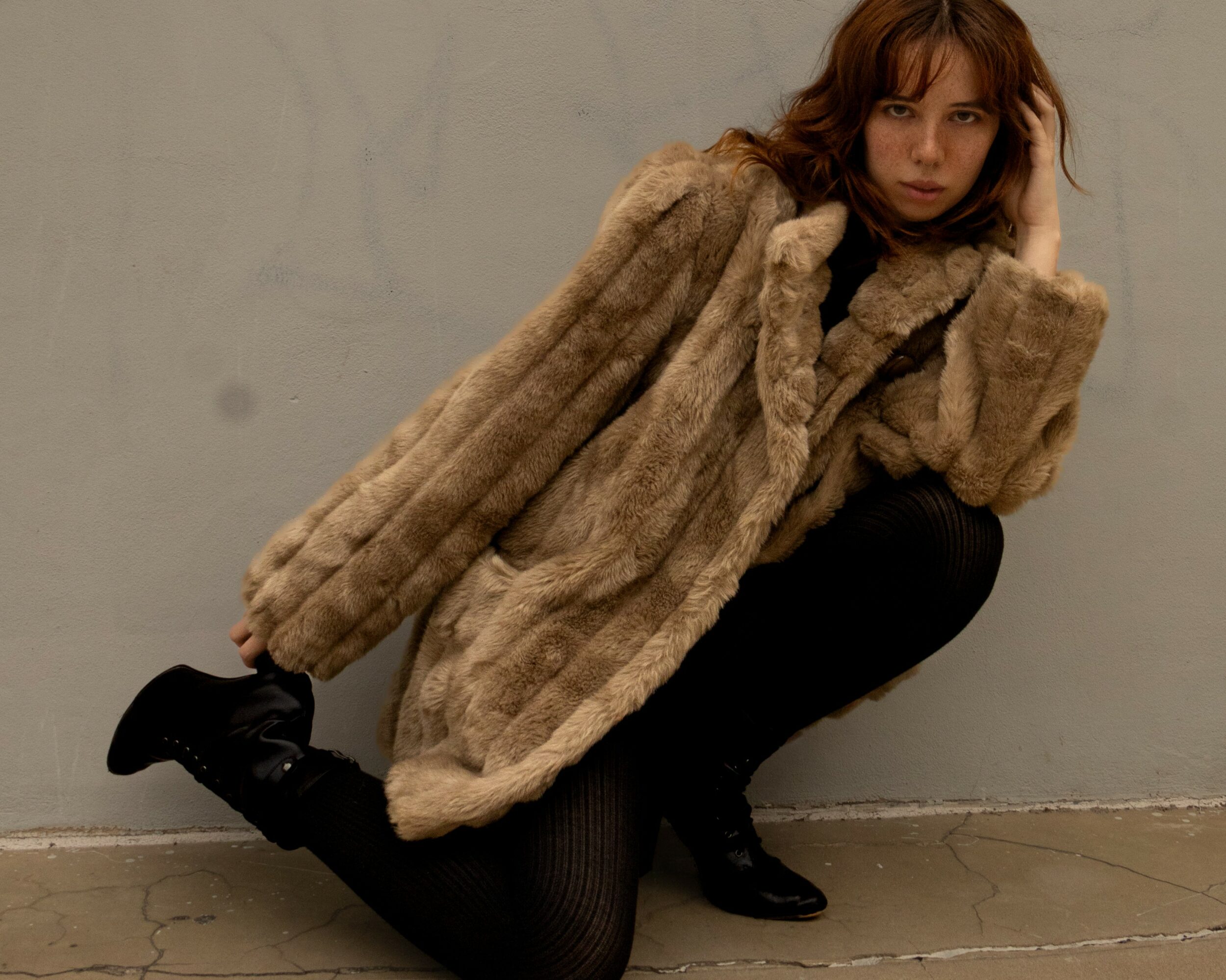 Person crouching against a plain wall, wearing a fur coat and black pants, with one hand on their head.