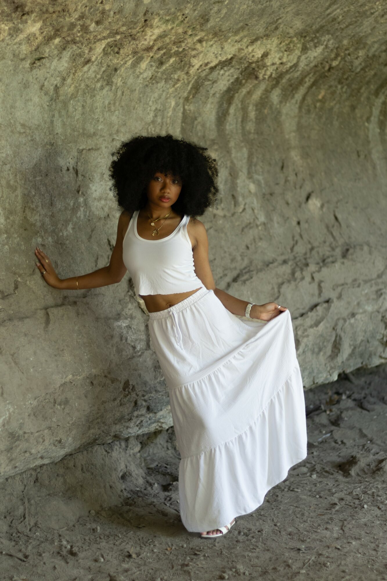 Woman with curly hair in a white tank top and long skirt standing against a textured wall.