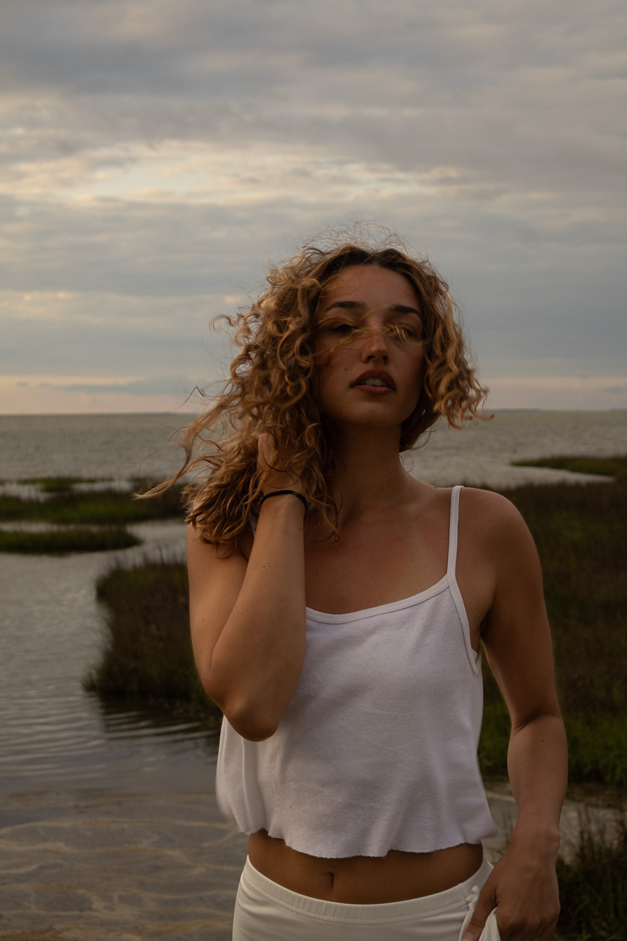 Young woman with curly hair standing outdoors near water, wearing a white tank top, looking at the camera.