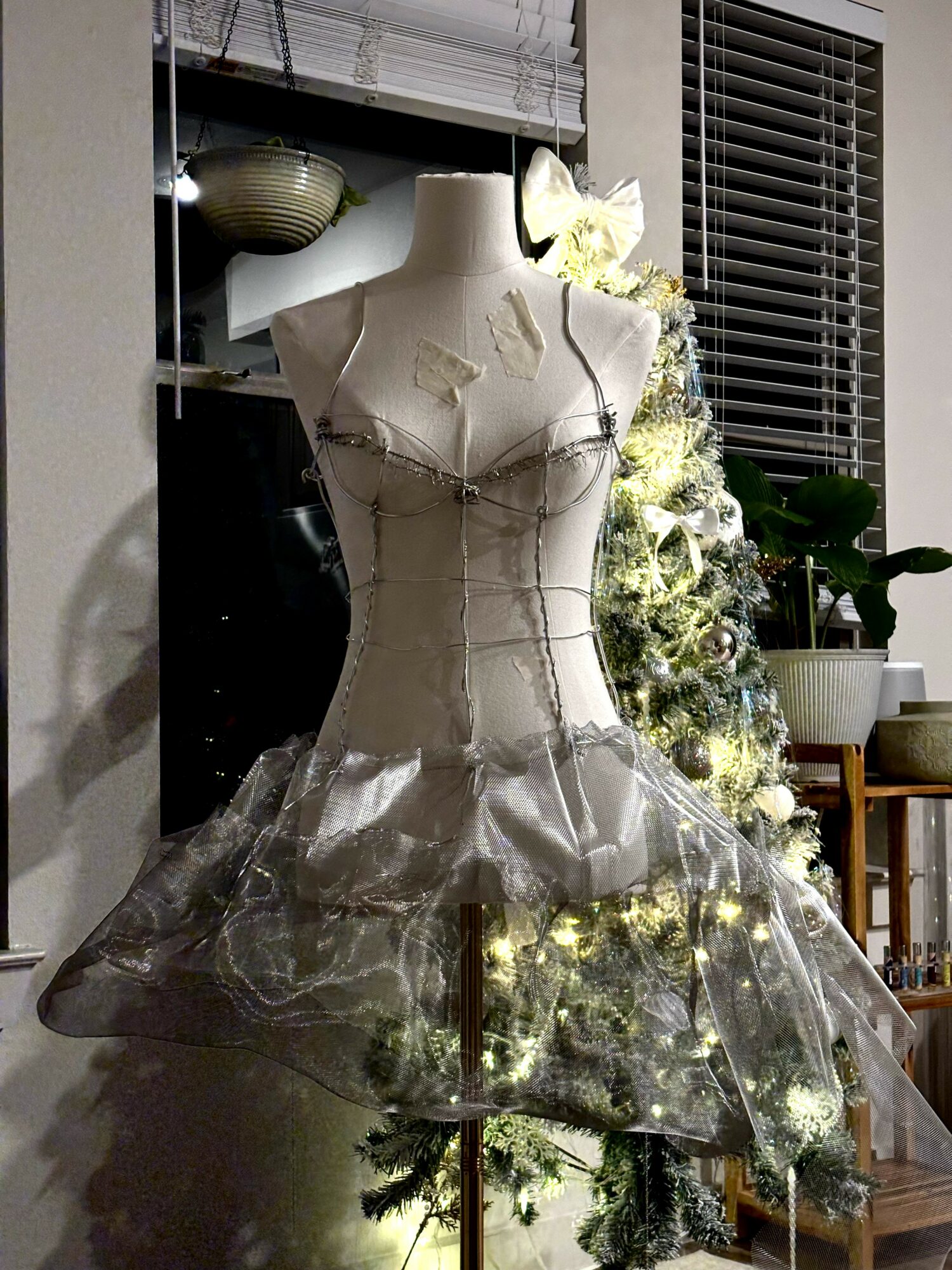 Mannequin torso dressed in a shiny, metallic dress with a fitted bodice and flared skirt, in a room with a decorated Christmas tree.
