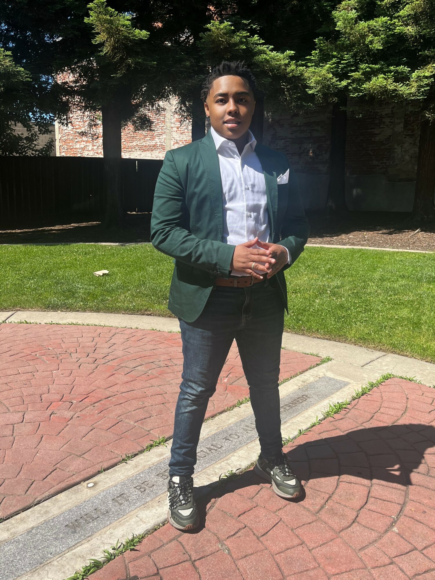 Young man in a green blazer and white shirt standing outdoors on a brick path, with trees and a fence in background.