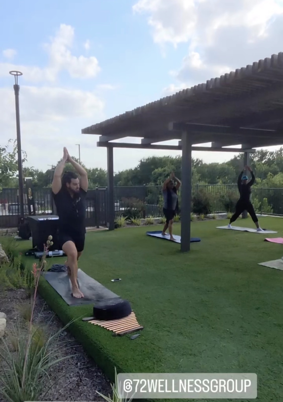 Three people practicing yoga outdoors on mats under a shelter, with a grassy area and trees in the background.