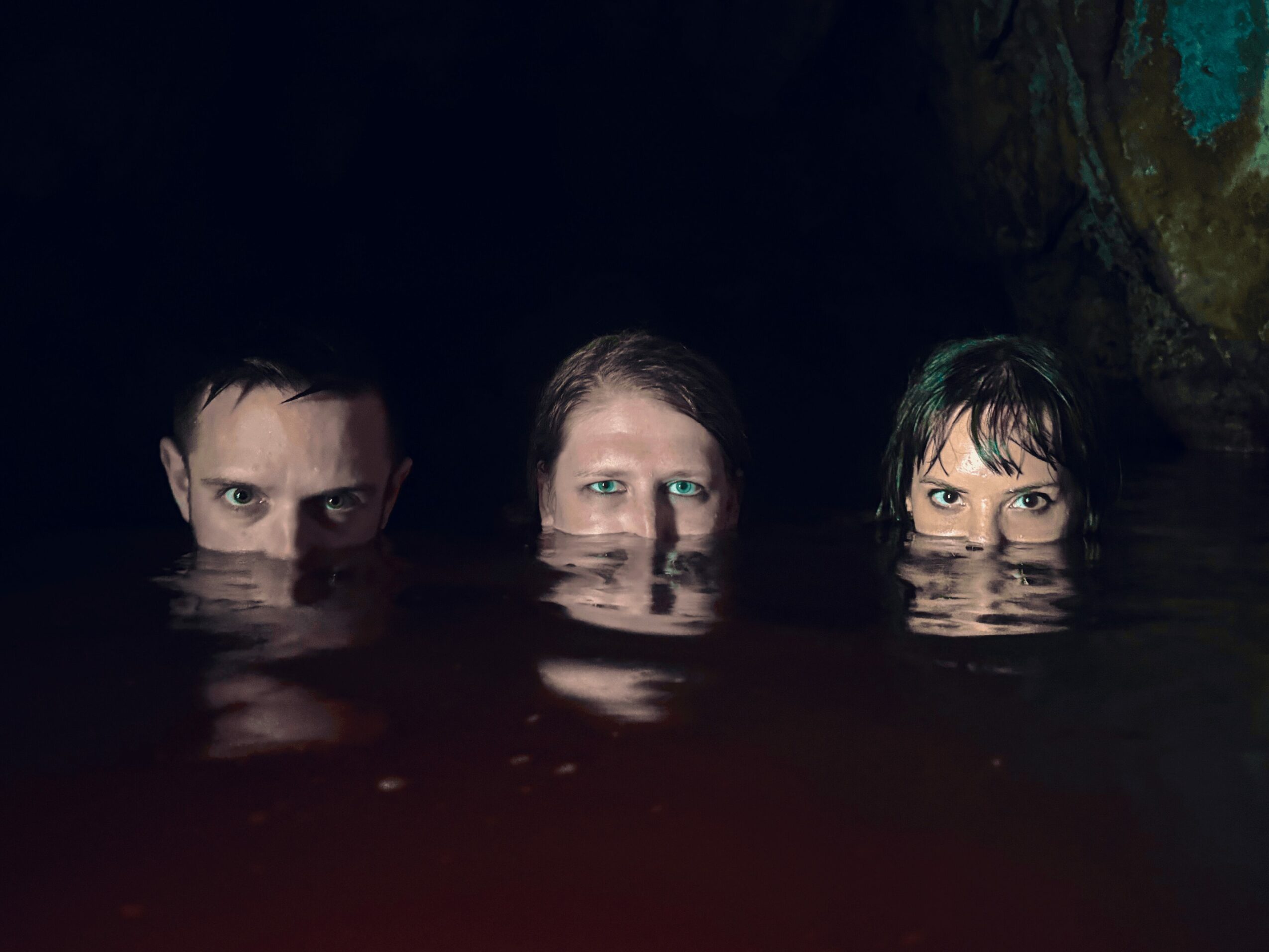 Three people partially submerged in water, with only their eyes and foreheads visible, in a dark environment.