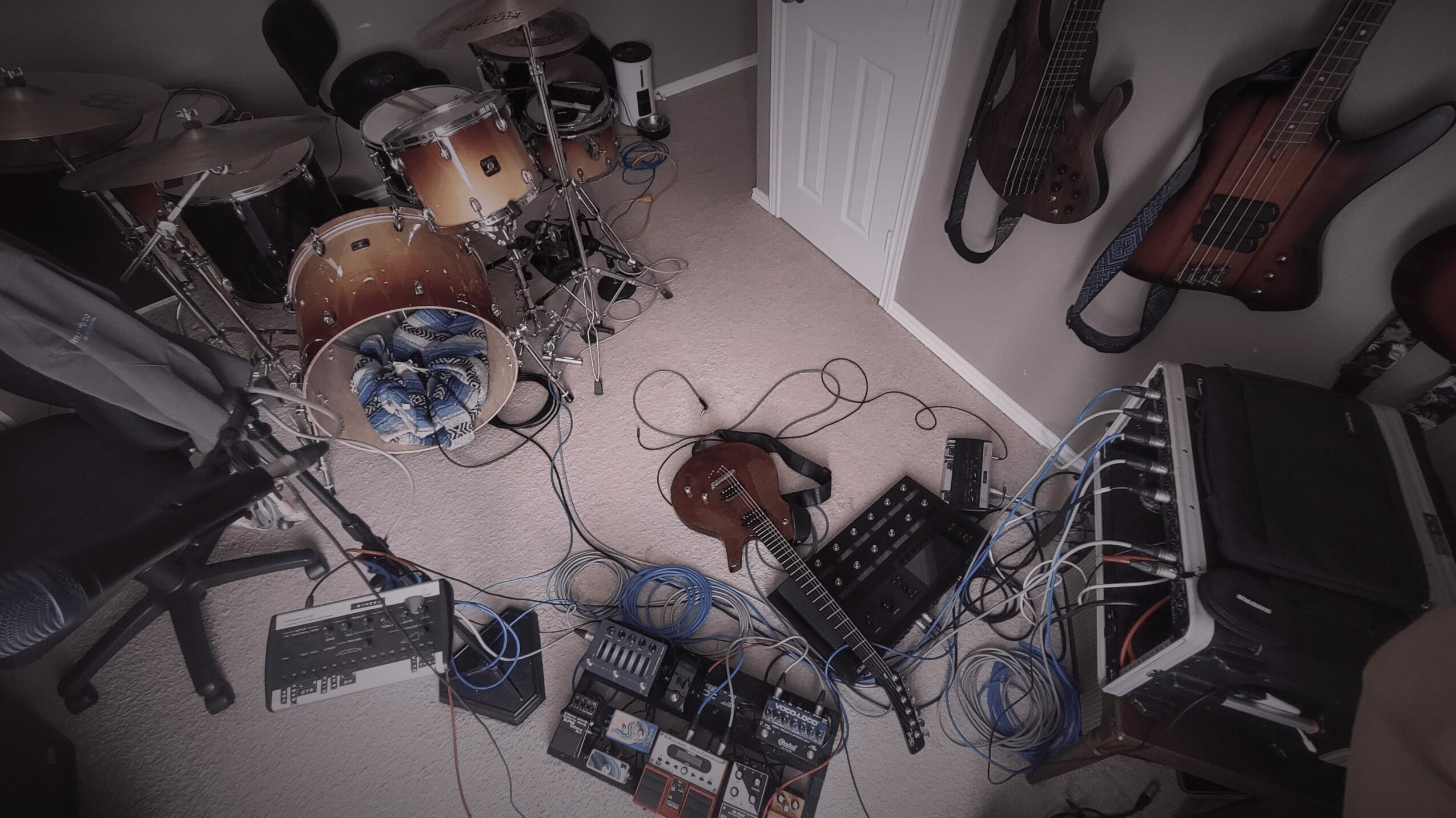 Musical instruments and equipment scattered on a carpeted floor, including drums, guitars, and audio gear, with cables tangled.