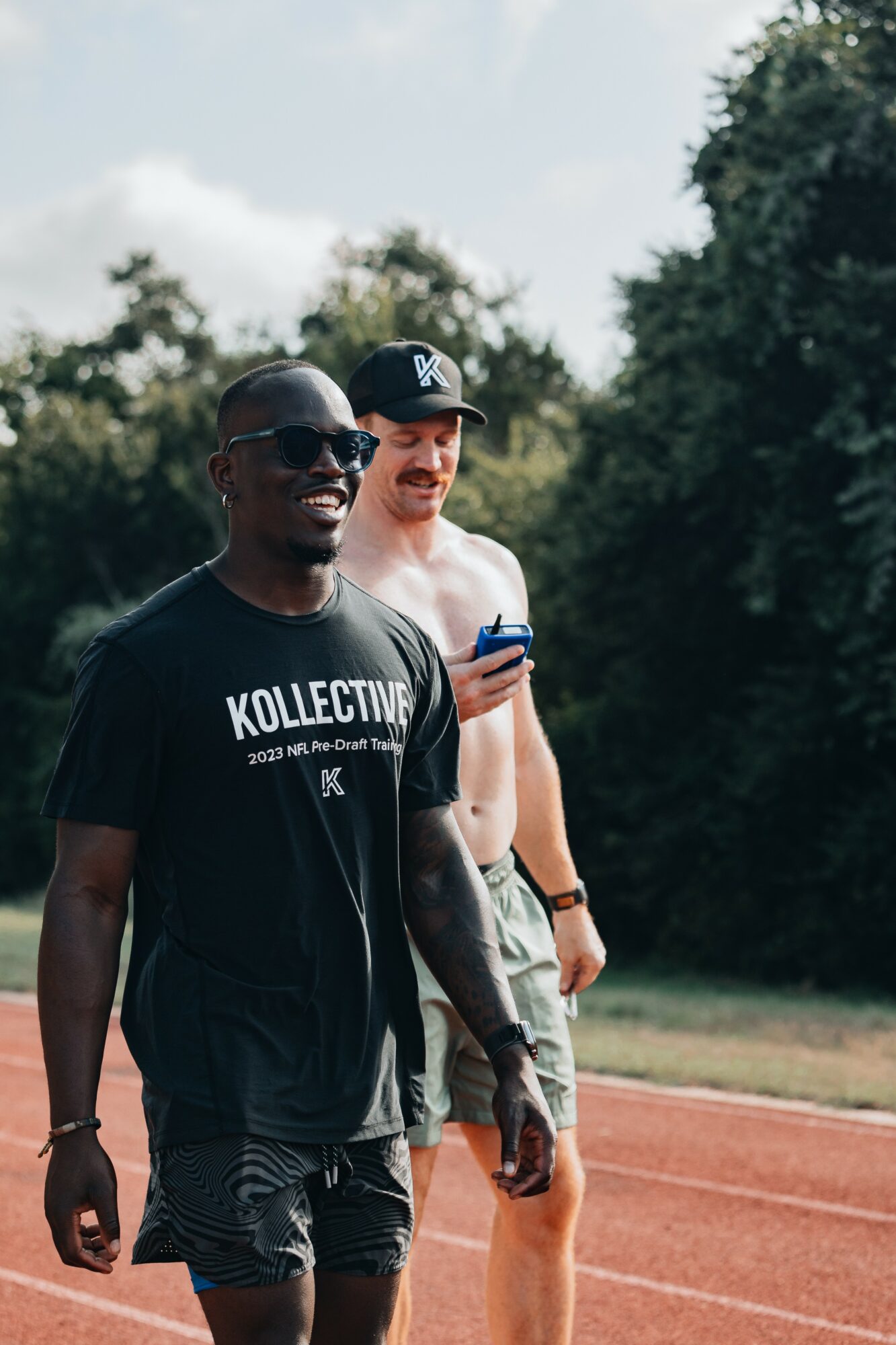 Two men walking outdoors on a track, one wearing sunglasses and a black T-shirt, the other in a cap and shirt, holding a phone.