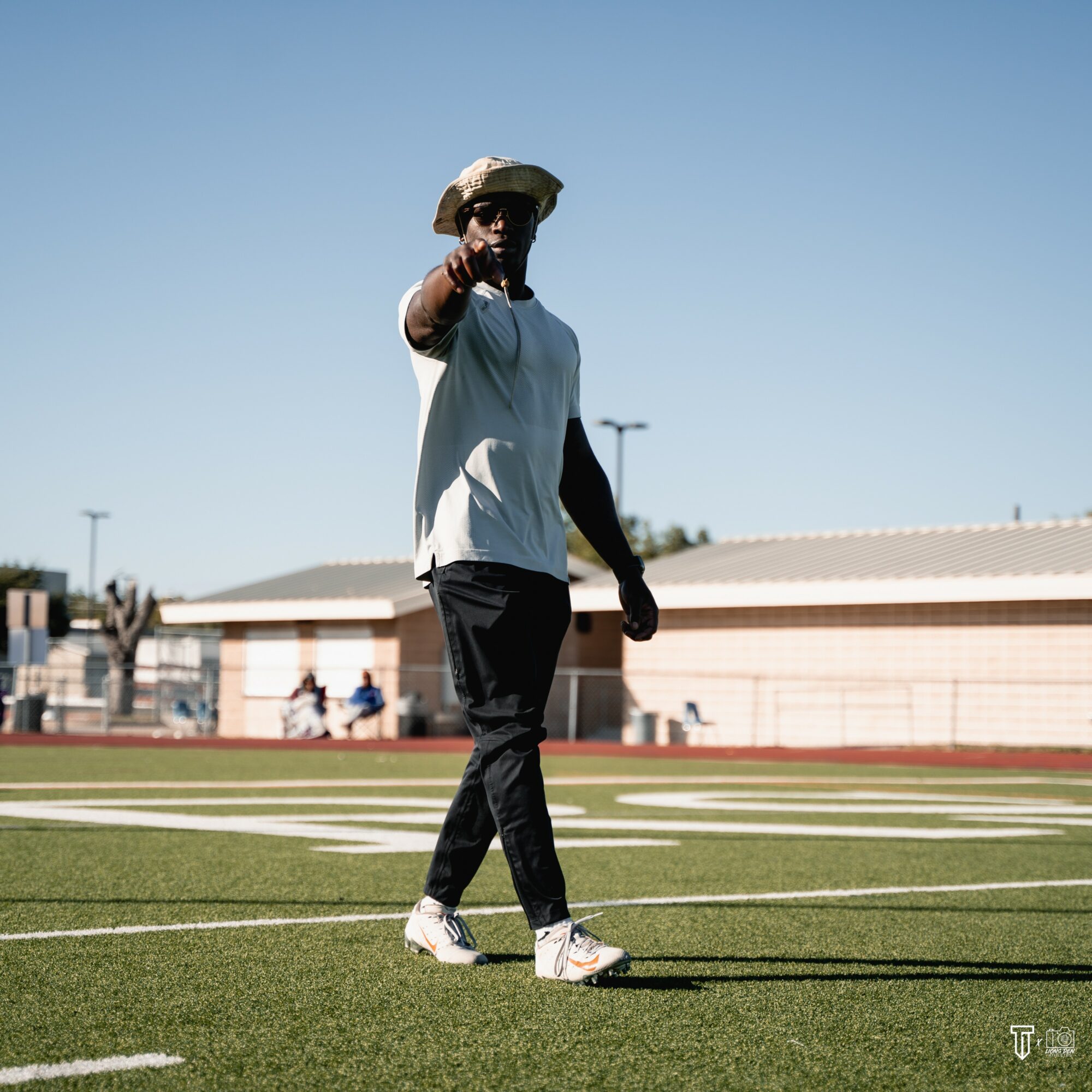 Person standing on a sports field pointing towards the camera, wearing a hat, sunglasses, and casual clothing, with a building and people in the background.