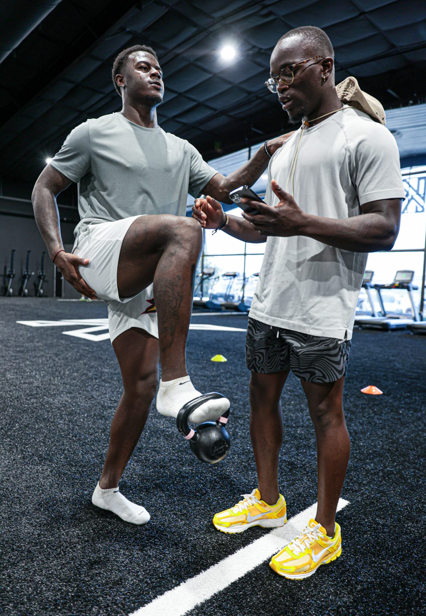 Two people in athletic clothing in a gym, one with a leg on a ball, the other holding a phone, discussing.