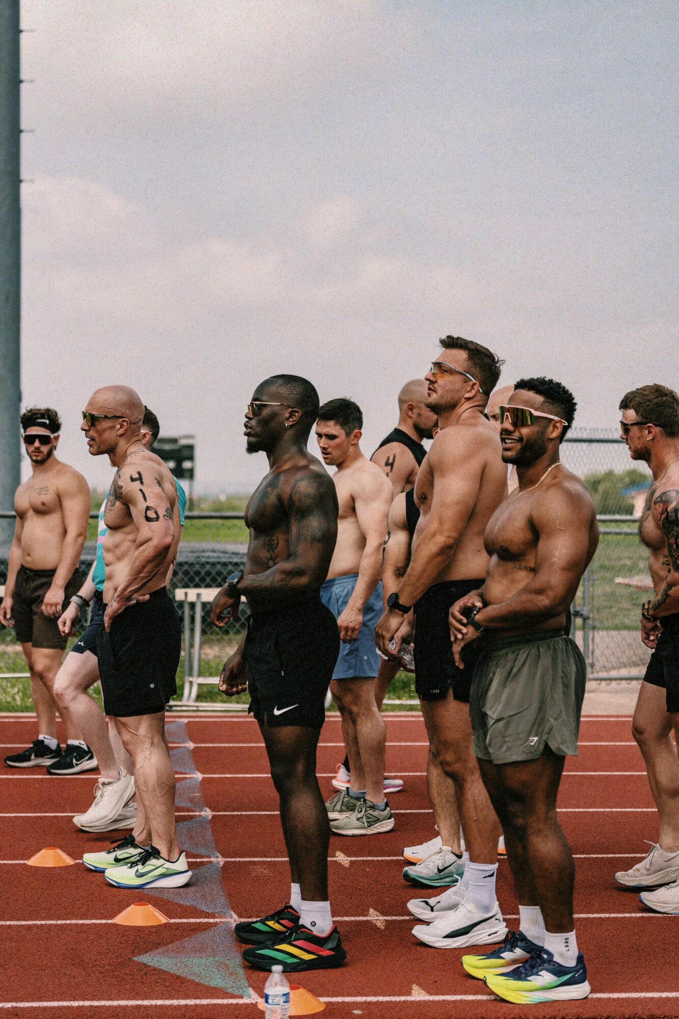 Group of shirtless men standing on a running track outdoors, some wearing sunglasses and athletic shoes, with a cloudy sky above.