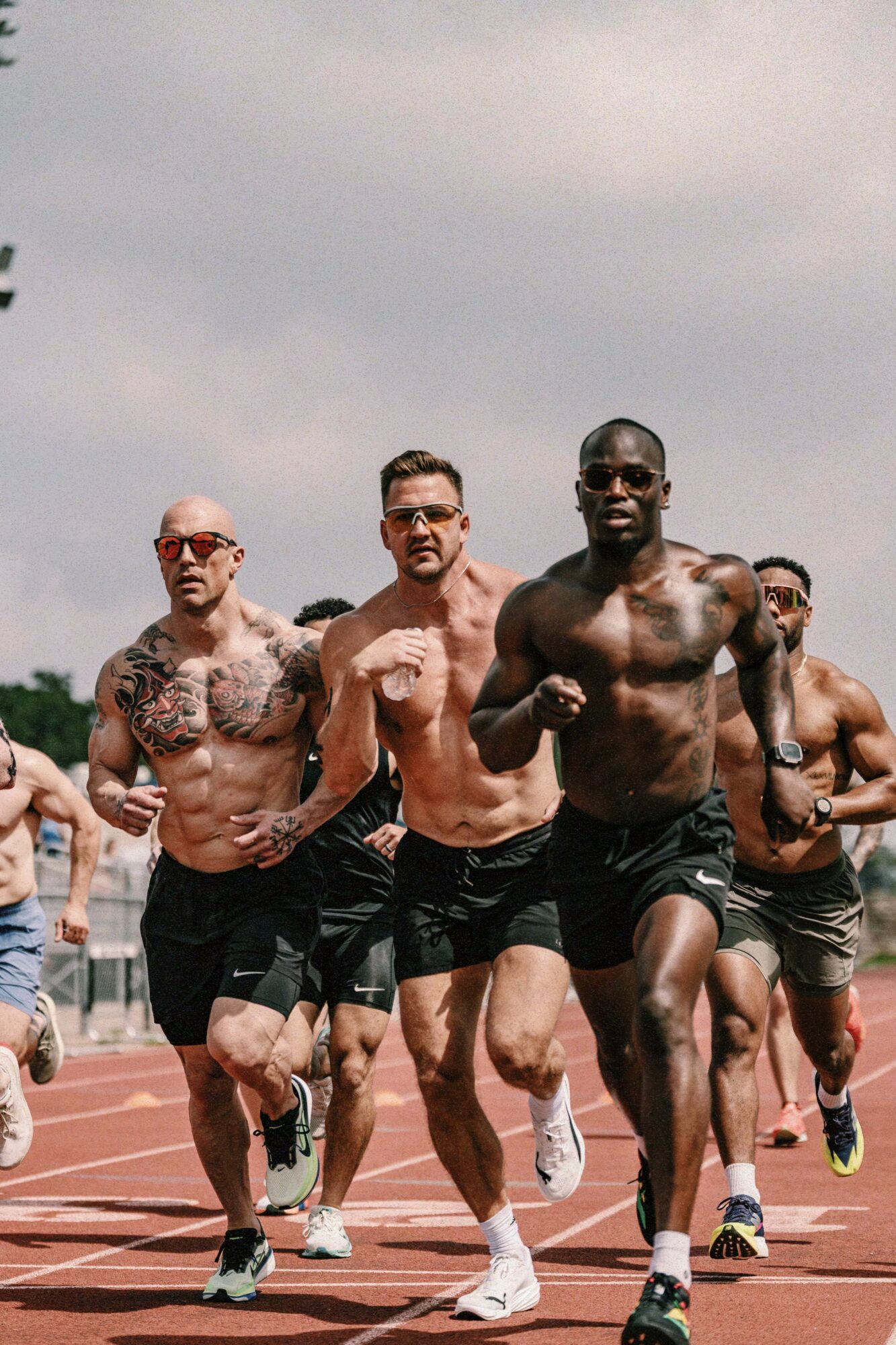 Group of shirtless male runners sprinting on a track outdoors under cloudy sky.