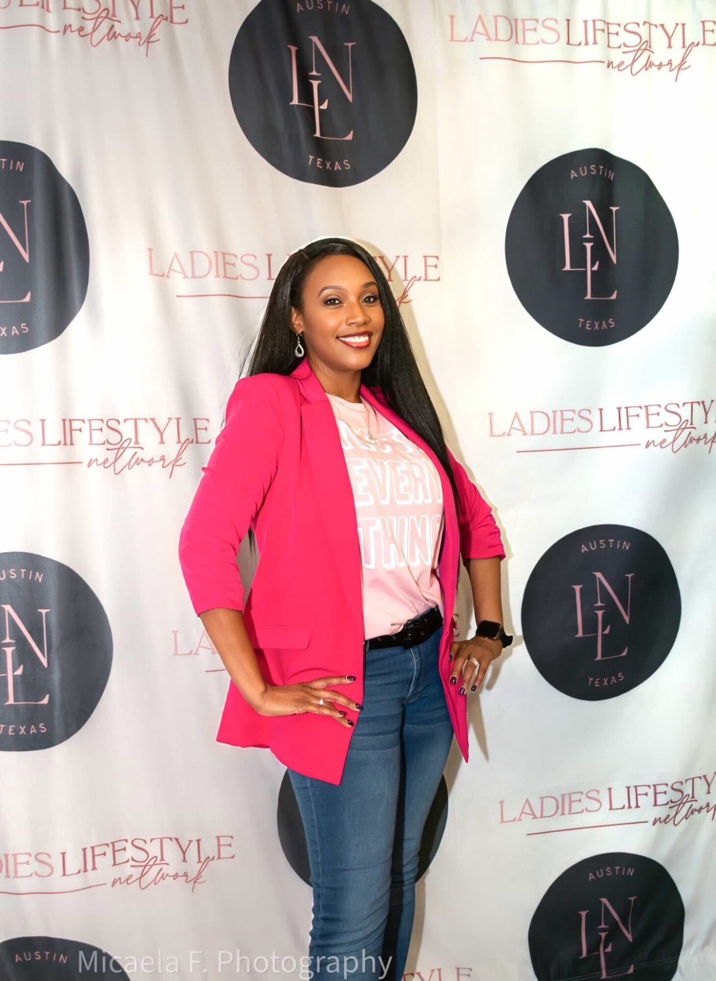 Woman in pink blazer and blue jeans smiling, standing in front of a backdrop with logos and text.