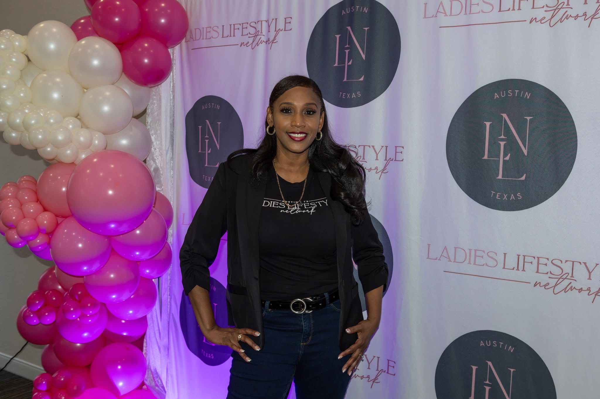 Woman standing in front of a backdrop with balloons on the left, smiling, wearing a black jacket and jeans.
