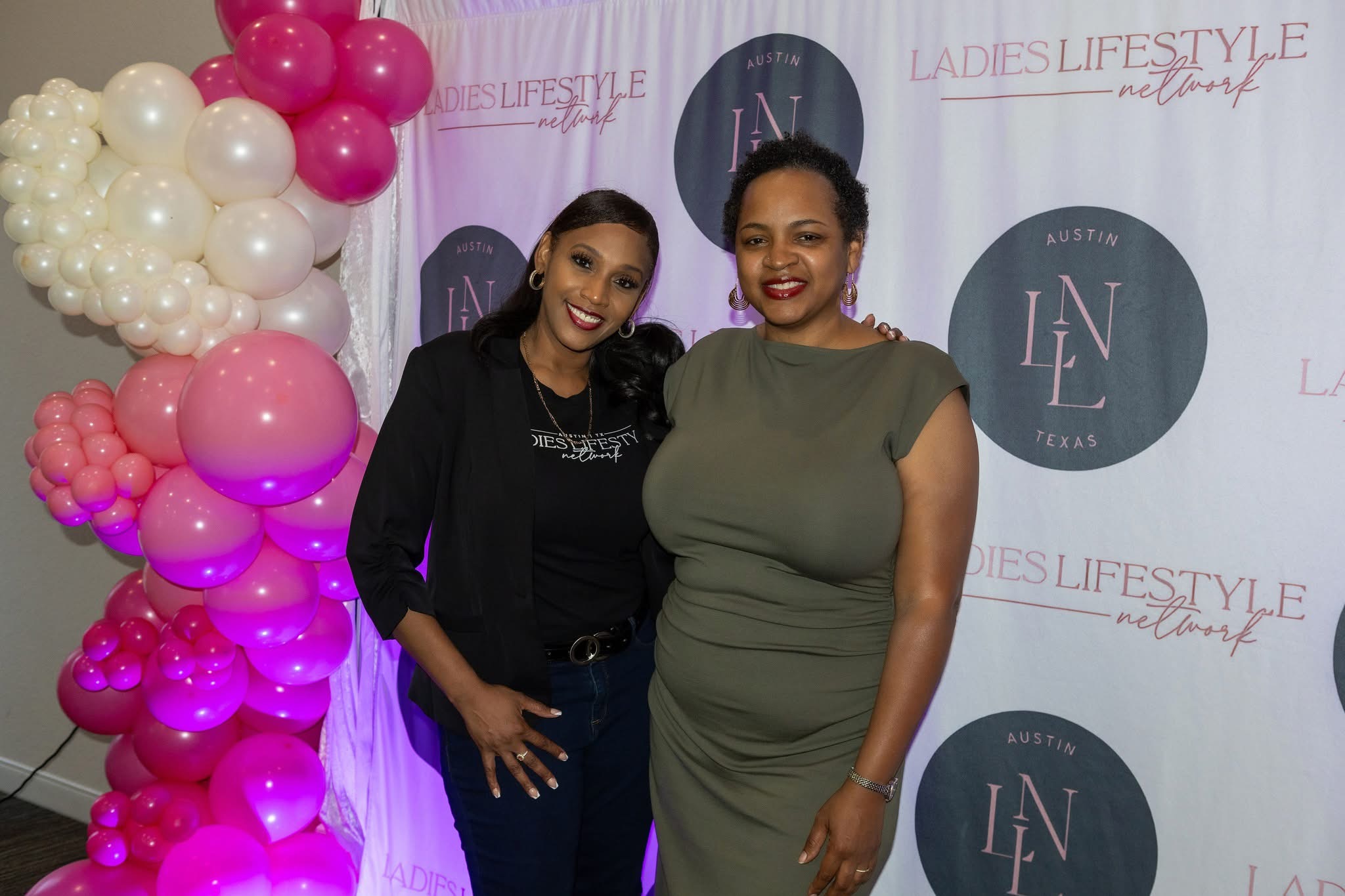Two women standing in front of a backdrop with balloons on the left side, smiling at the camera.